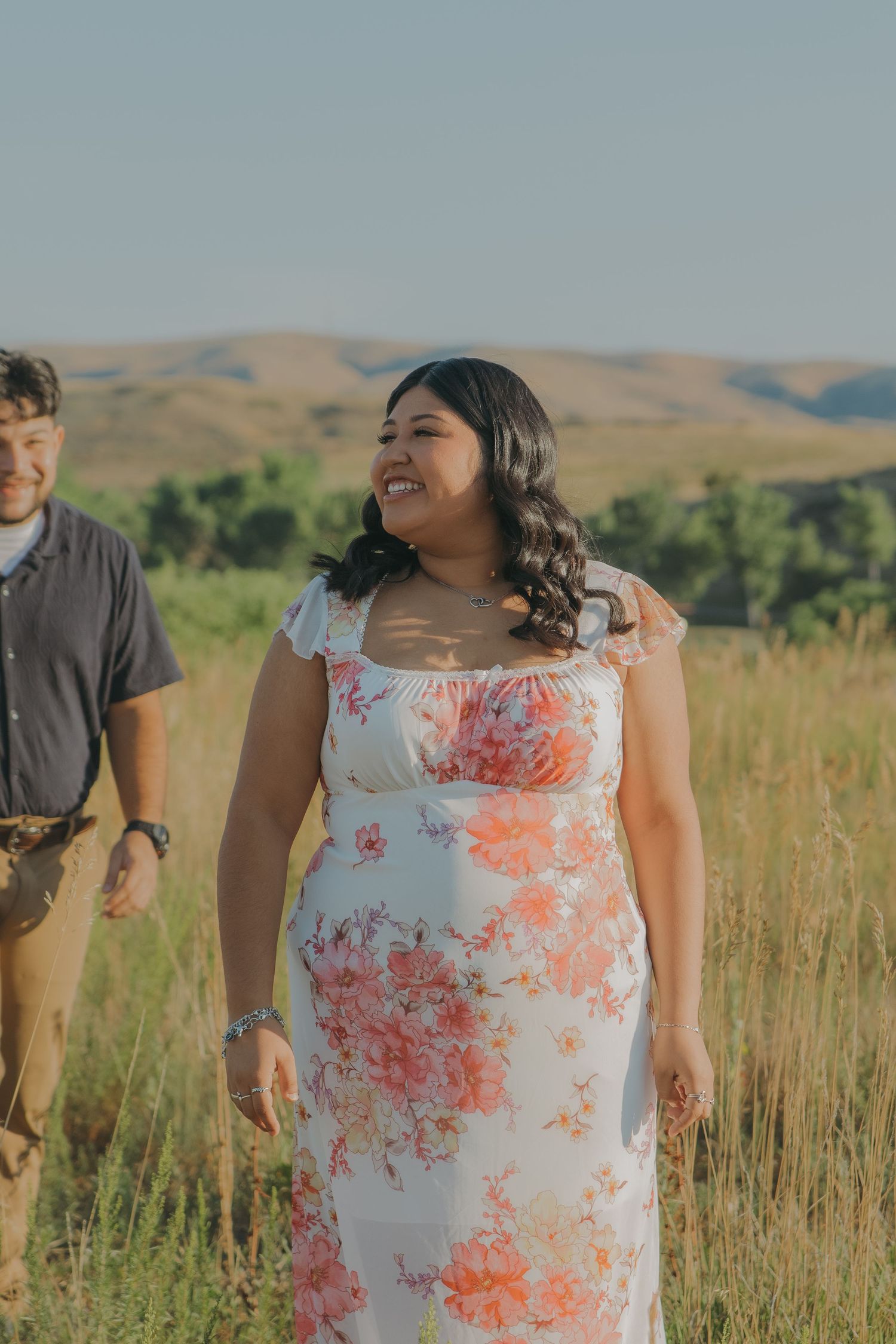 A woman in a floral dress laughs joyfully against a backdrop of golden hills and blue sky.