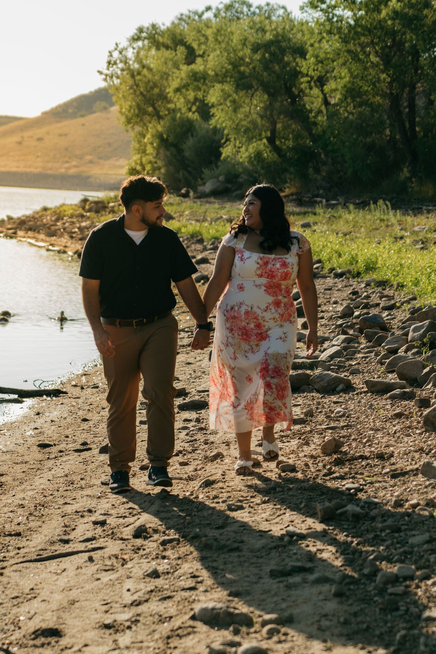 A couple walks along a rocky shoreline with trees and hills in the background.