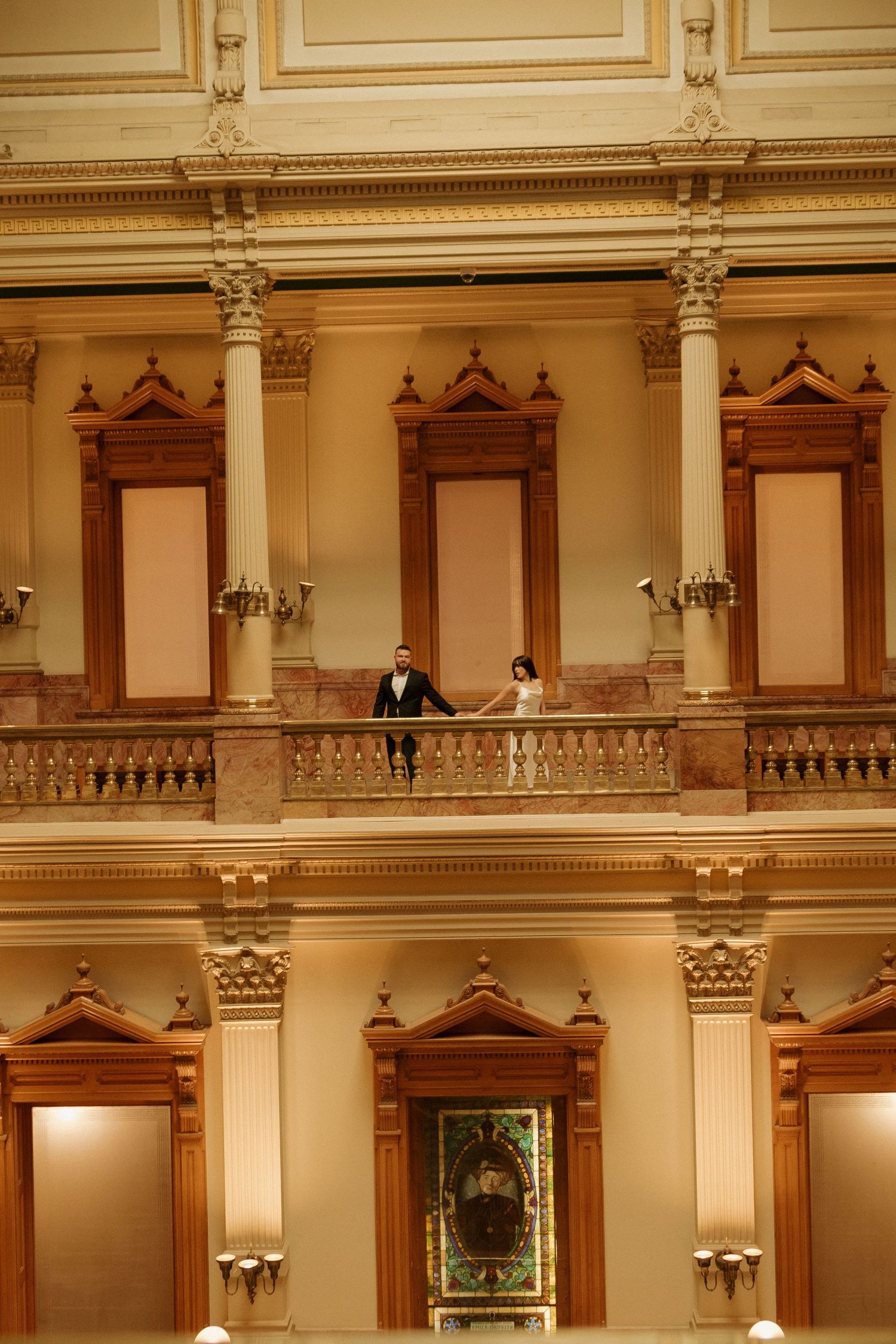 Couple poses on ornate balcony of historic building with classical architecture.