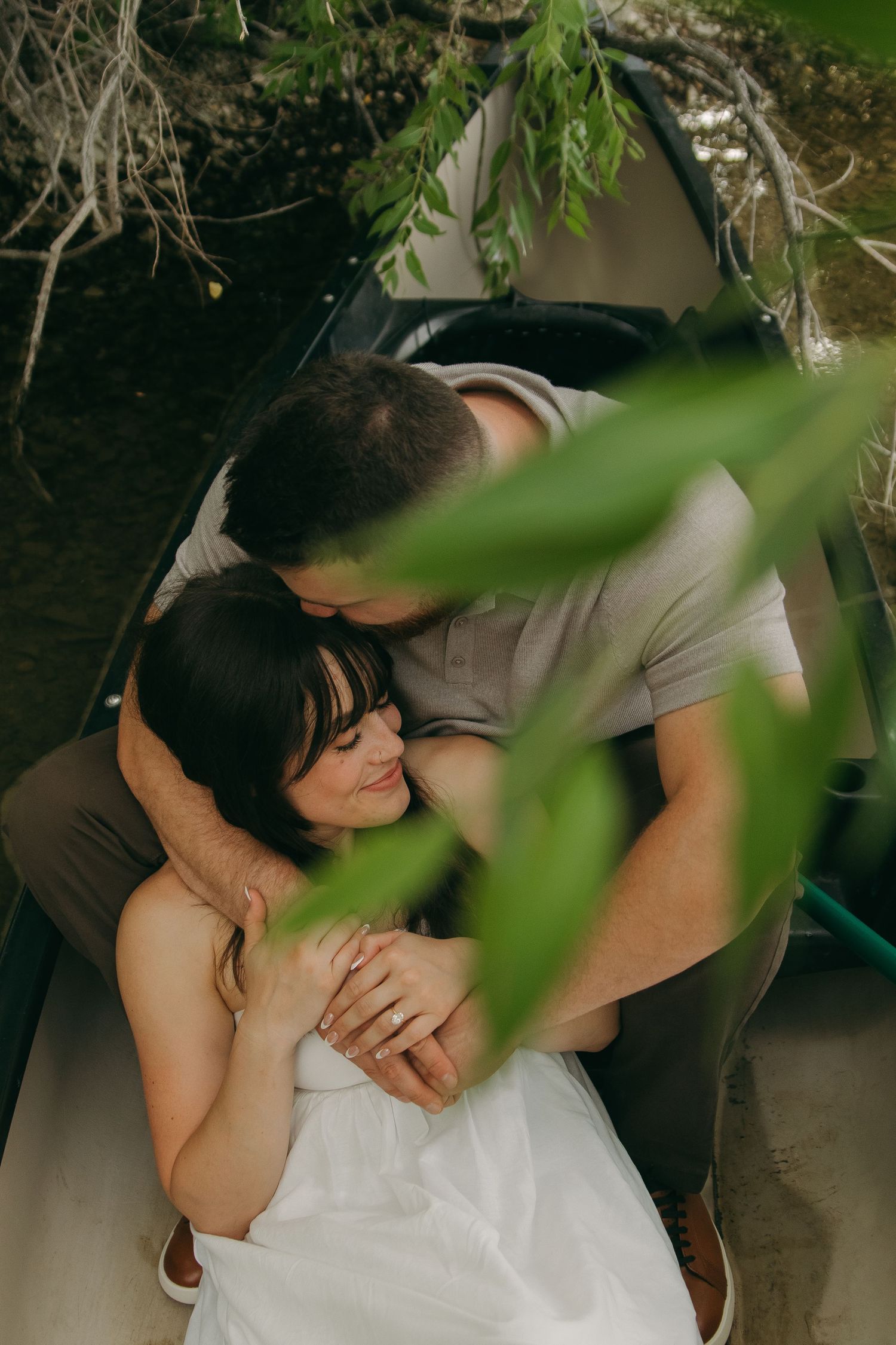 A couple shares a romantic embrace in a canoe framed by green leaves.