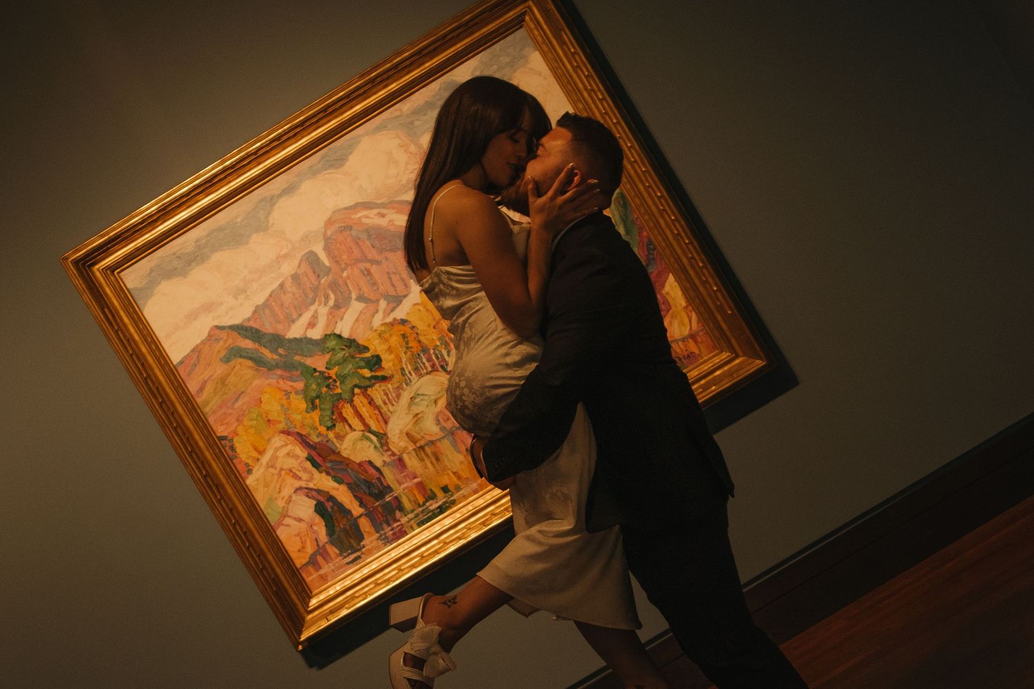 Two people view paintings in a dimly lit museum gallery with golden frames on display.
