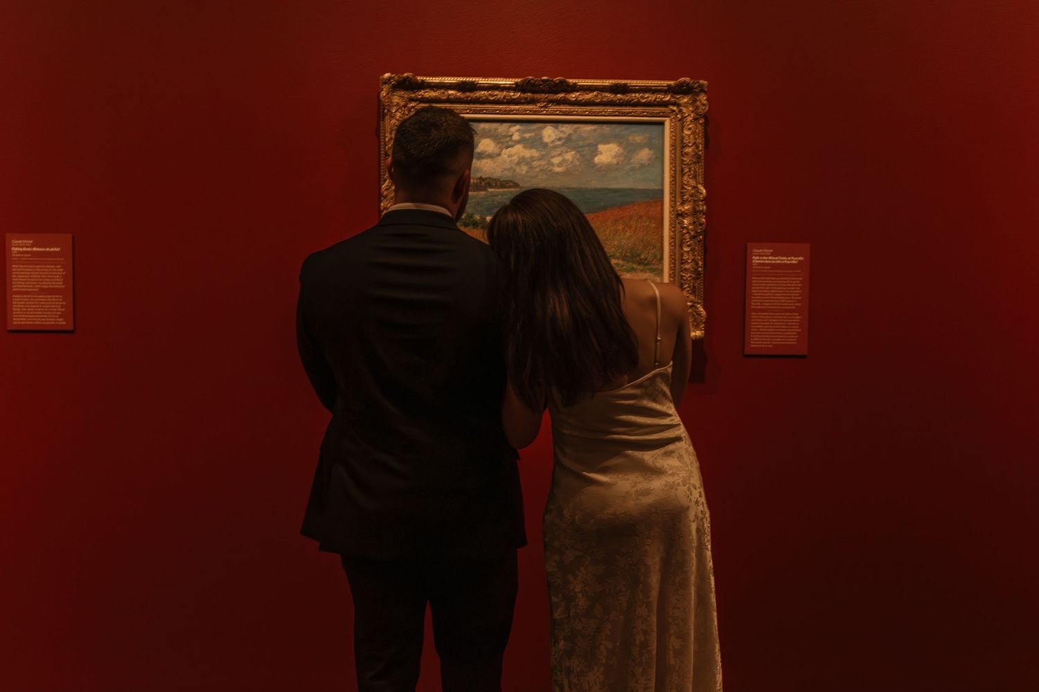 Gallery visitors viewing artwork displayed on deep red walls in low lighting.