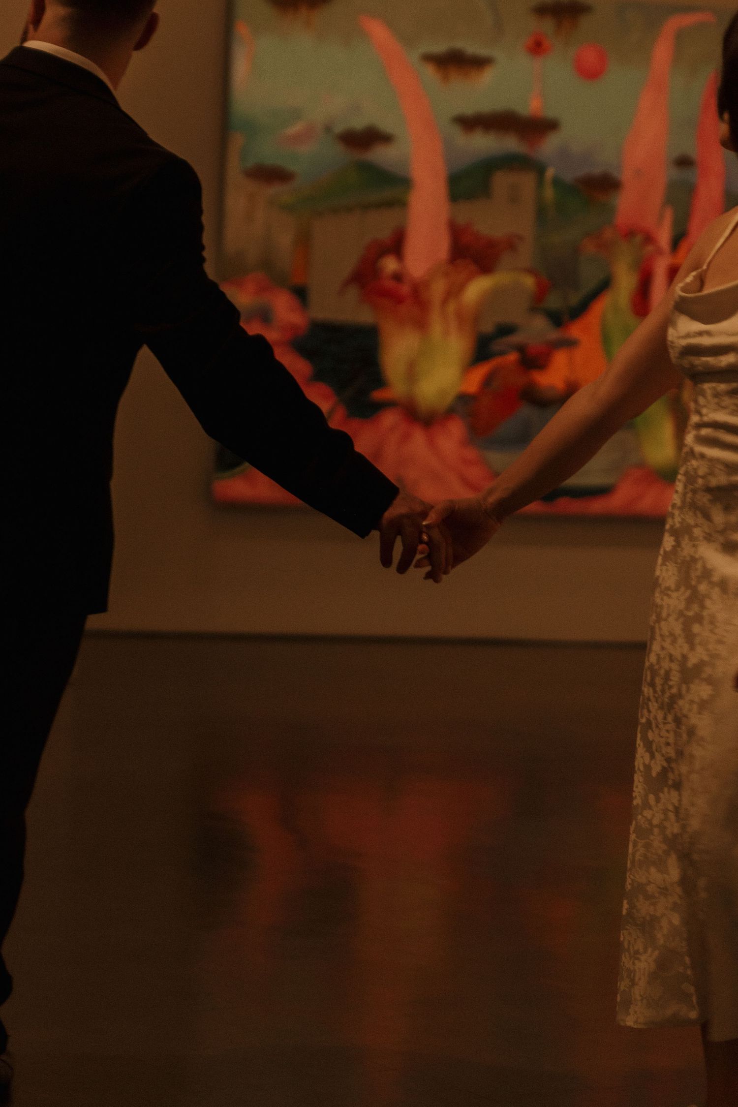 A couple holds hands in front of vibrant abstract artwork in a dimly lit gallery.
