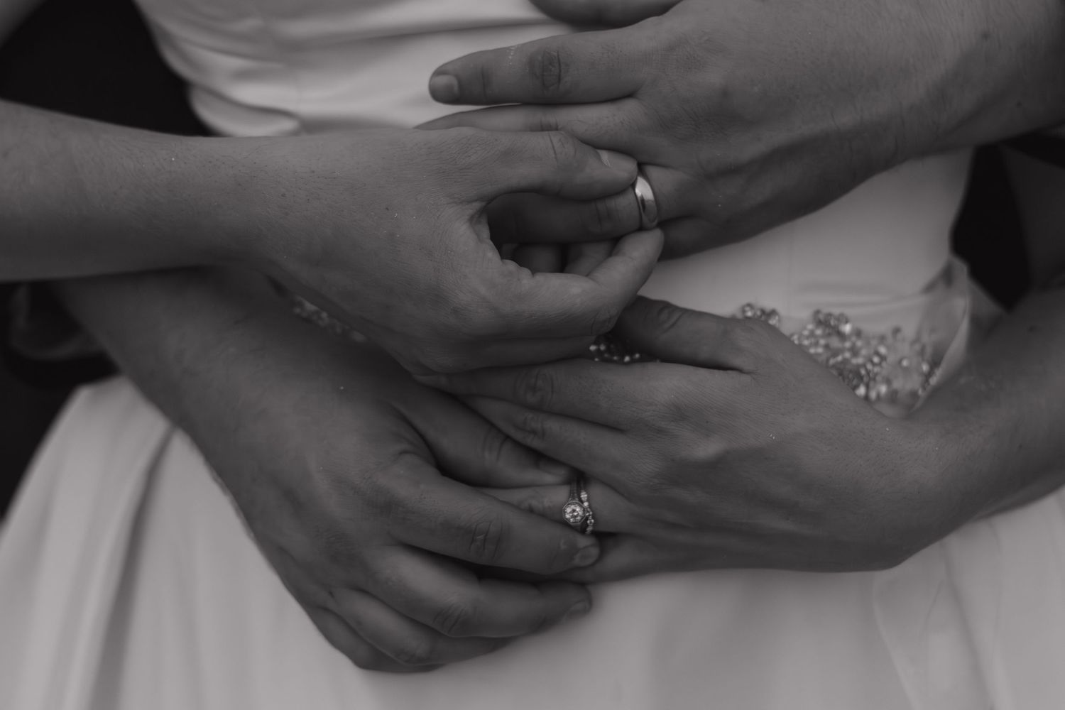 Close-up black and white photo of hands displaying wedding rings against a dress.
