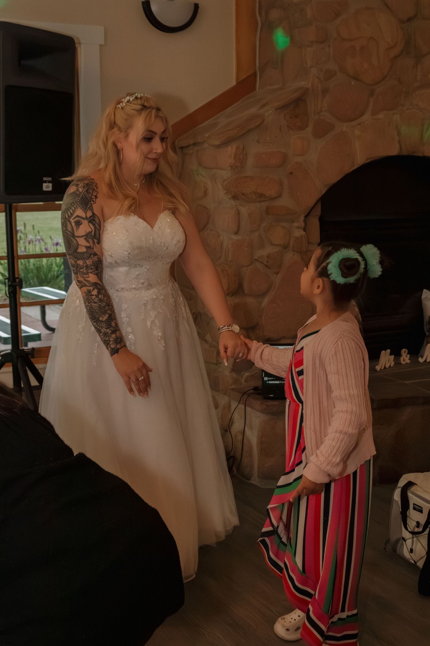A bride in a white gown interacting with young guest at wedding reception near stone fireplace.