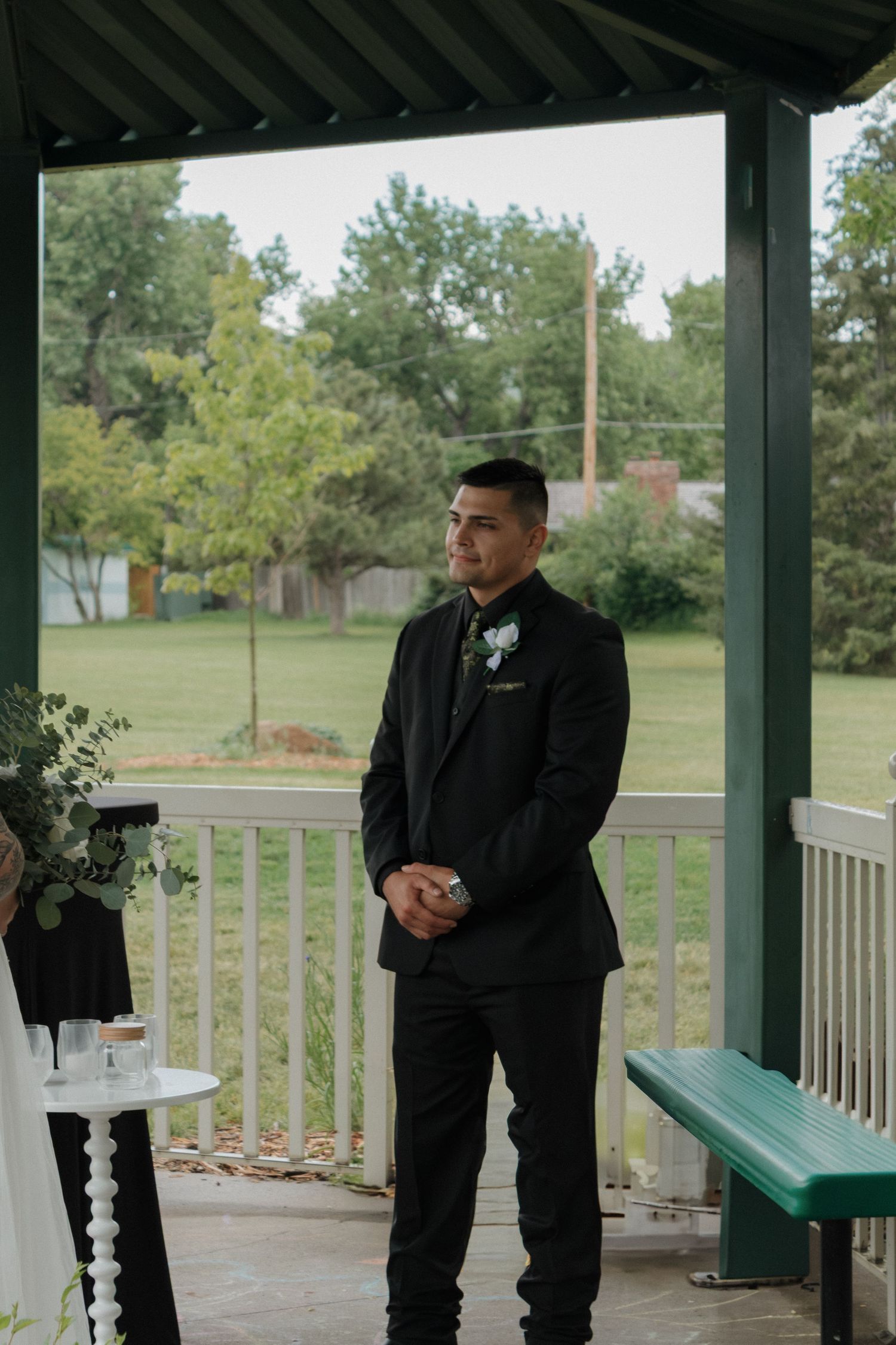 A groom in a black suit stands on a covered porch during a wedding ceremony.