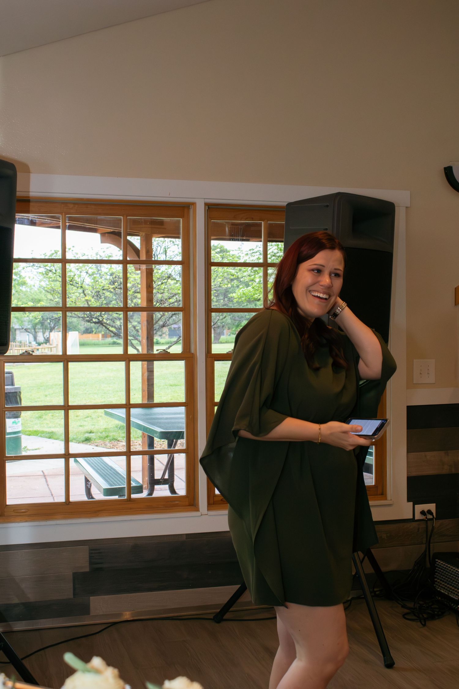 Woman in olive green dress speaking at event with windows and natural lighting.