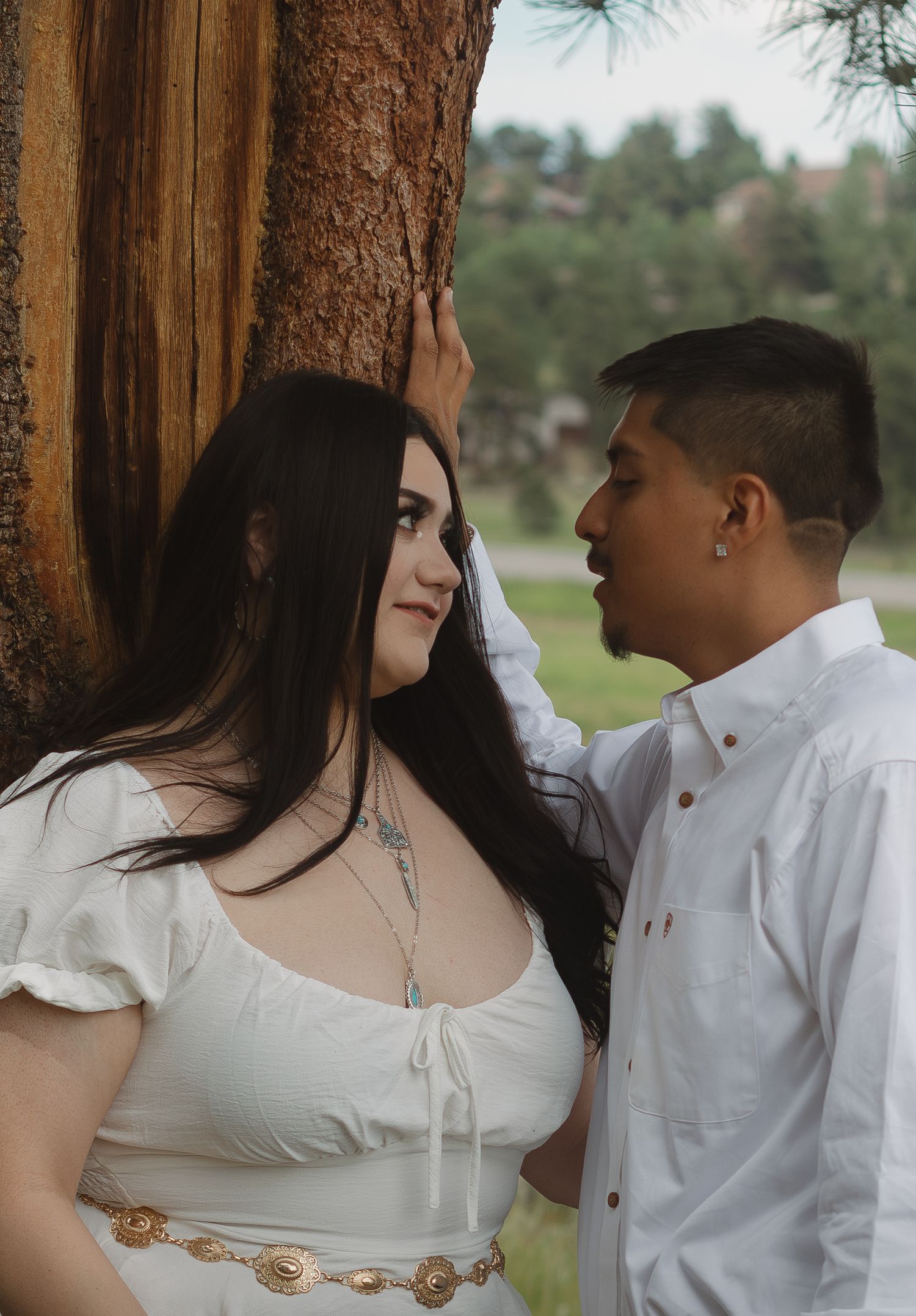 Romantic couple poses against a large tree trunk in a natural outdoor setting with mountains in background.