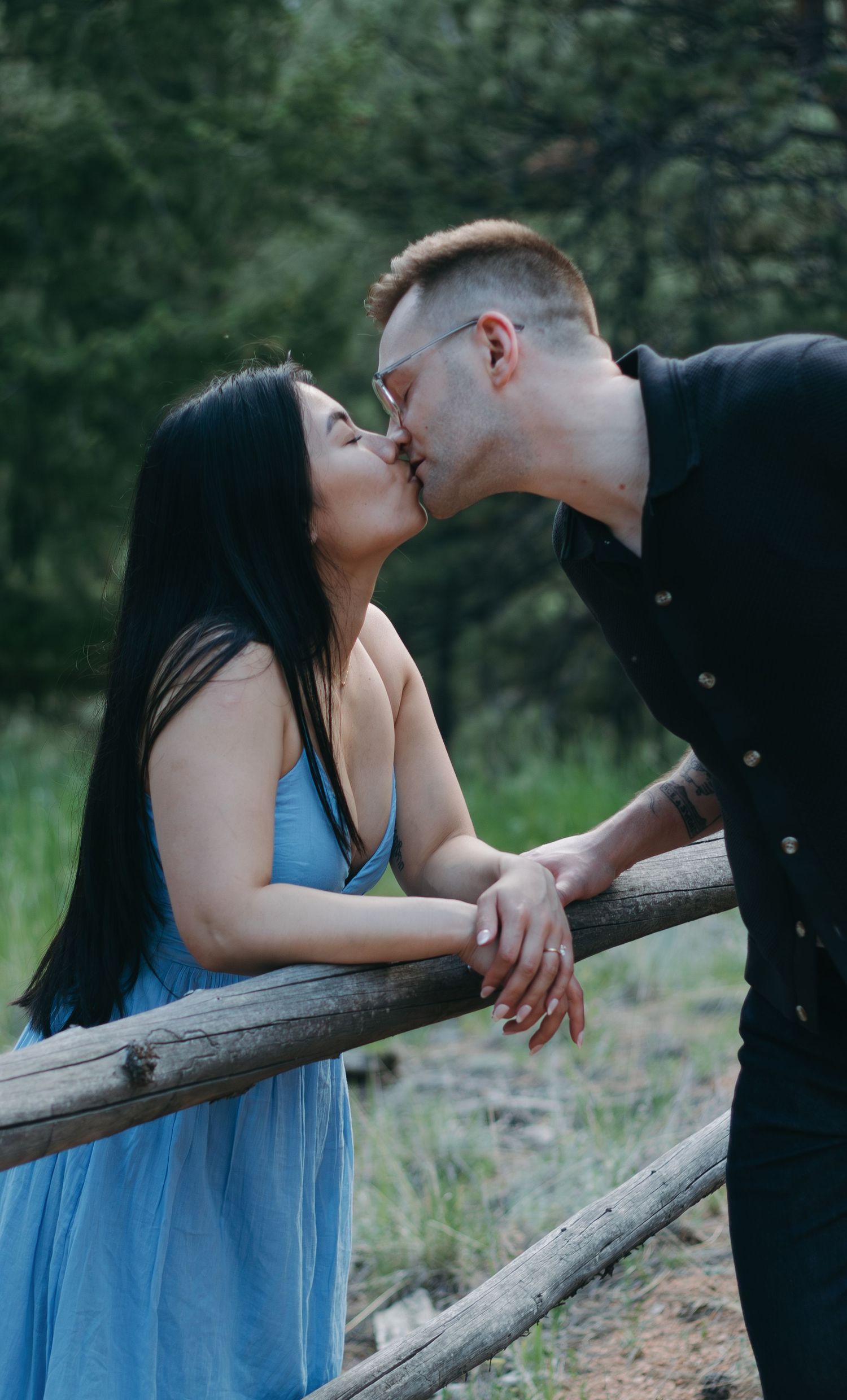 A romantic couple sharing a kiss while leaning against a wooden fence in a natural outdoor setting.