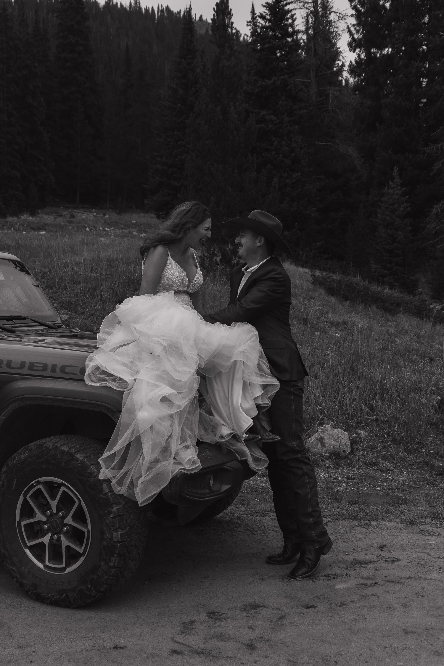 A black and white photo of a couple by their vehicle in a mountain setting.