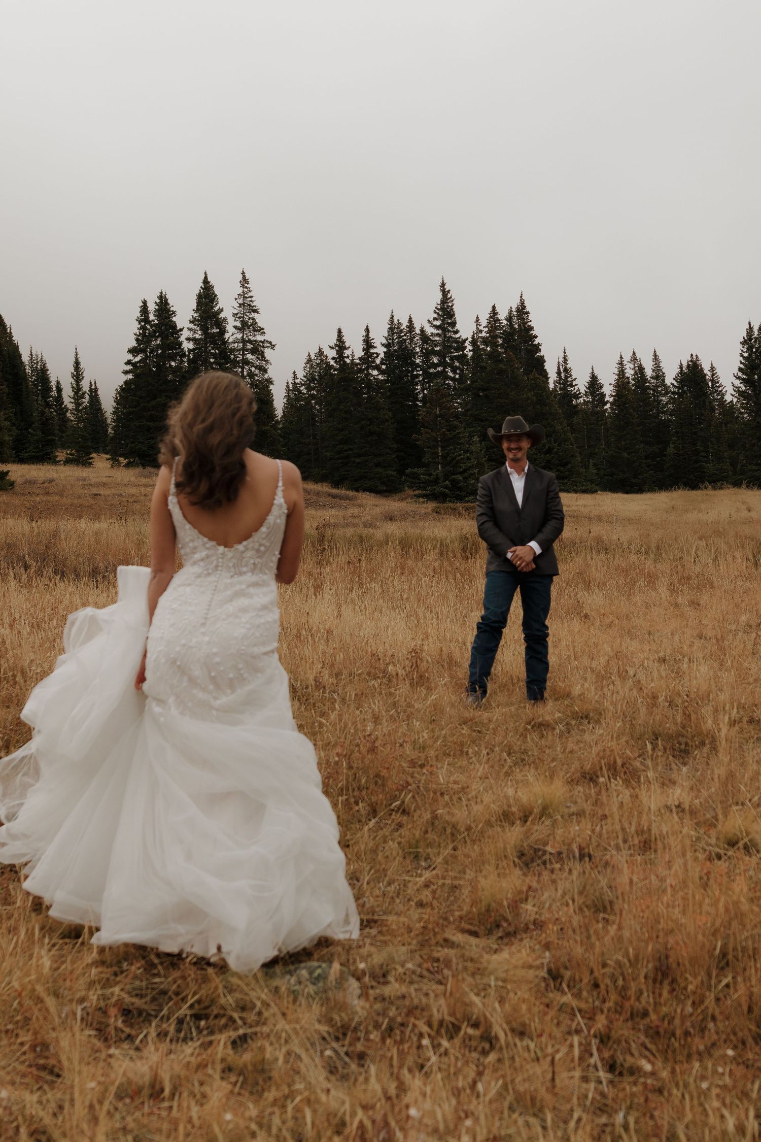 A couple in wedding attire playfully interacts in a golden mountain meadow surrounded by pine trees.