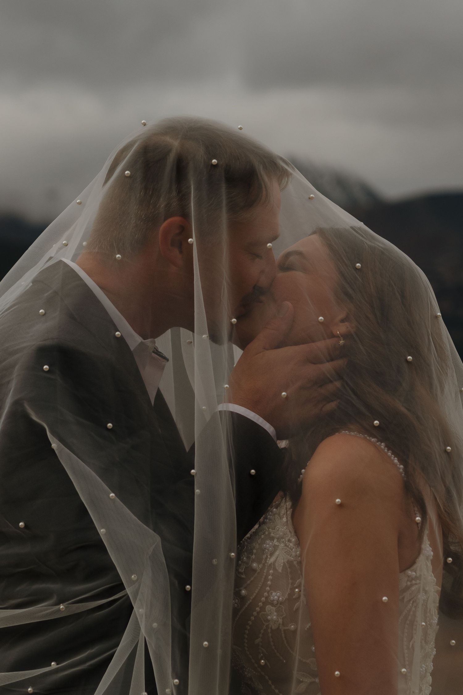 Romantic wedding moment under a delicate veil with mountains in the background.