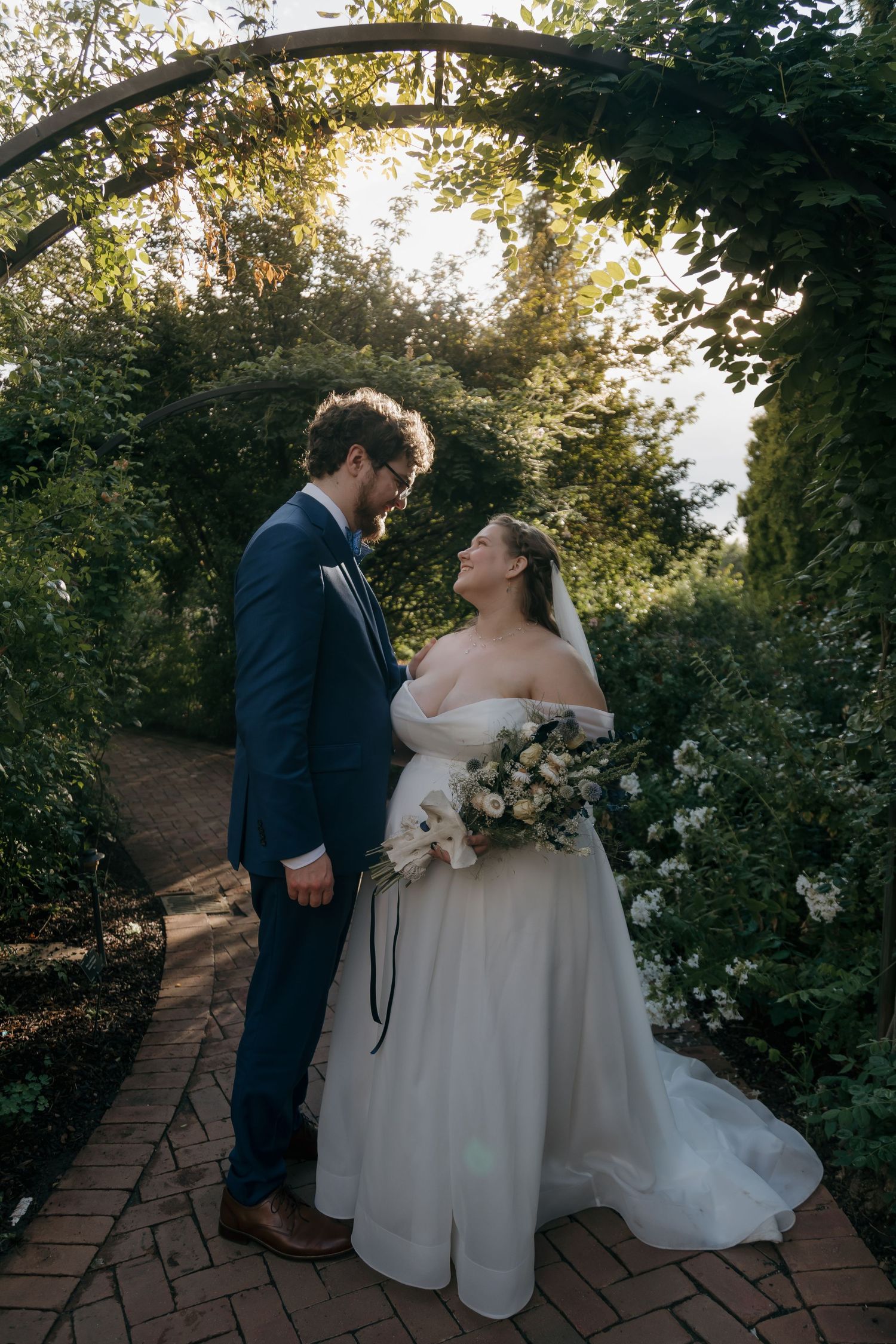 Bride and groom share a romantic moment under a garden arch, bride holding a white bouquet in golden sunlight.