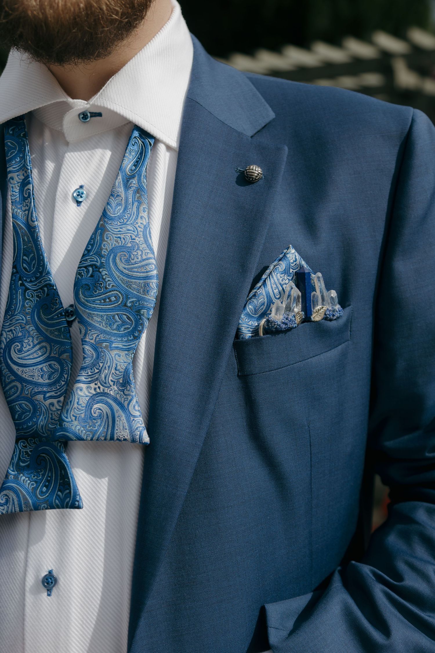 Close-up of groom in blue suit with paisley bow tie, matching vest, pocket square, and lapel pin.