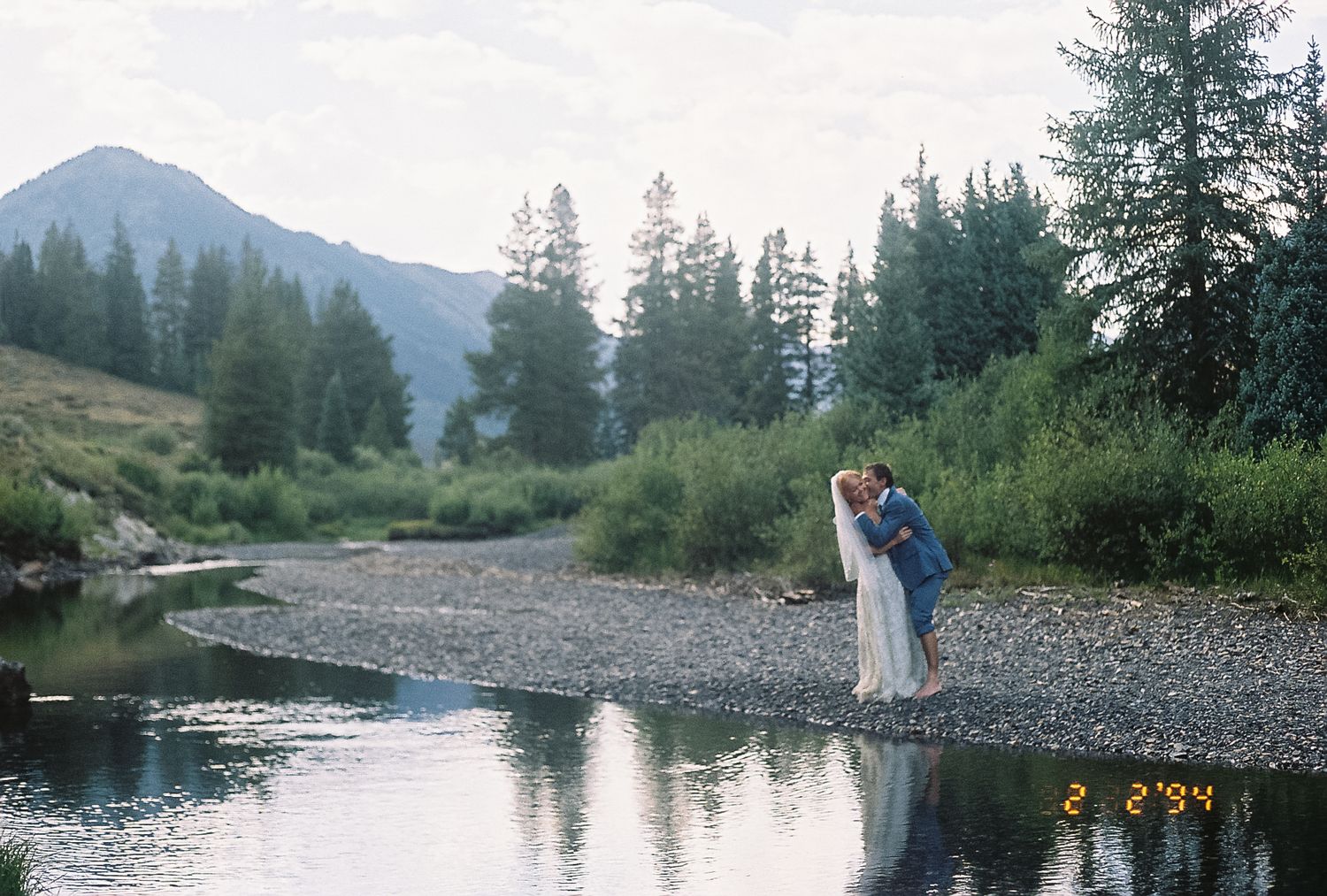 Bride and groom sharing a romantic kiss by a scenic mountain river on their wedding day.