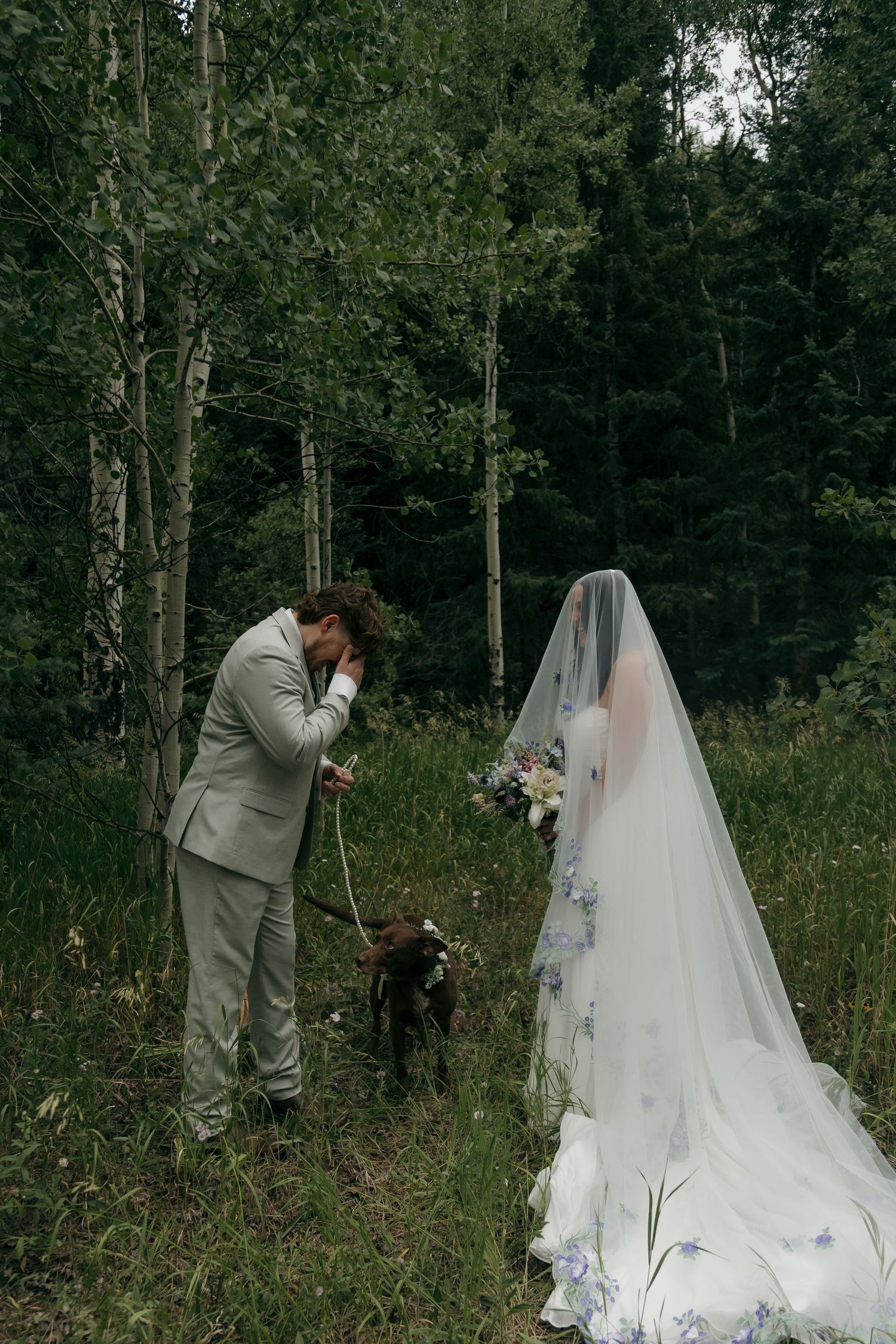 Emotional groom in sage suit bends to pet dog while bride holds wildflower bouquet in forest.