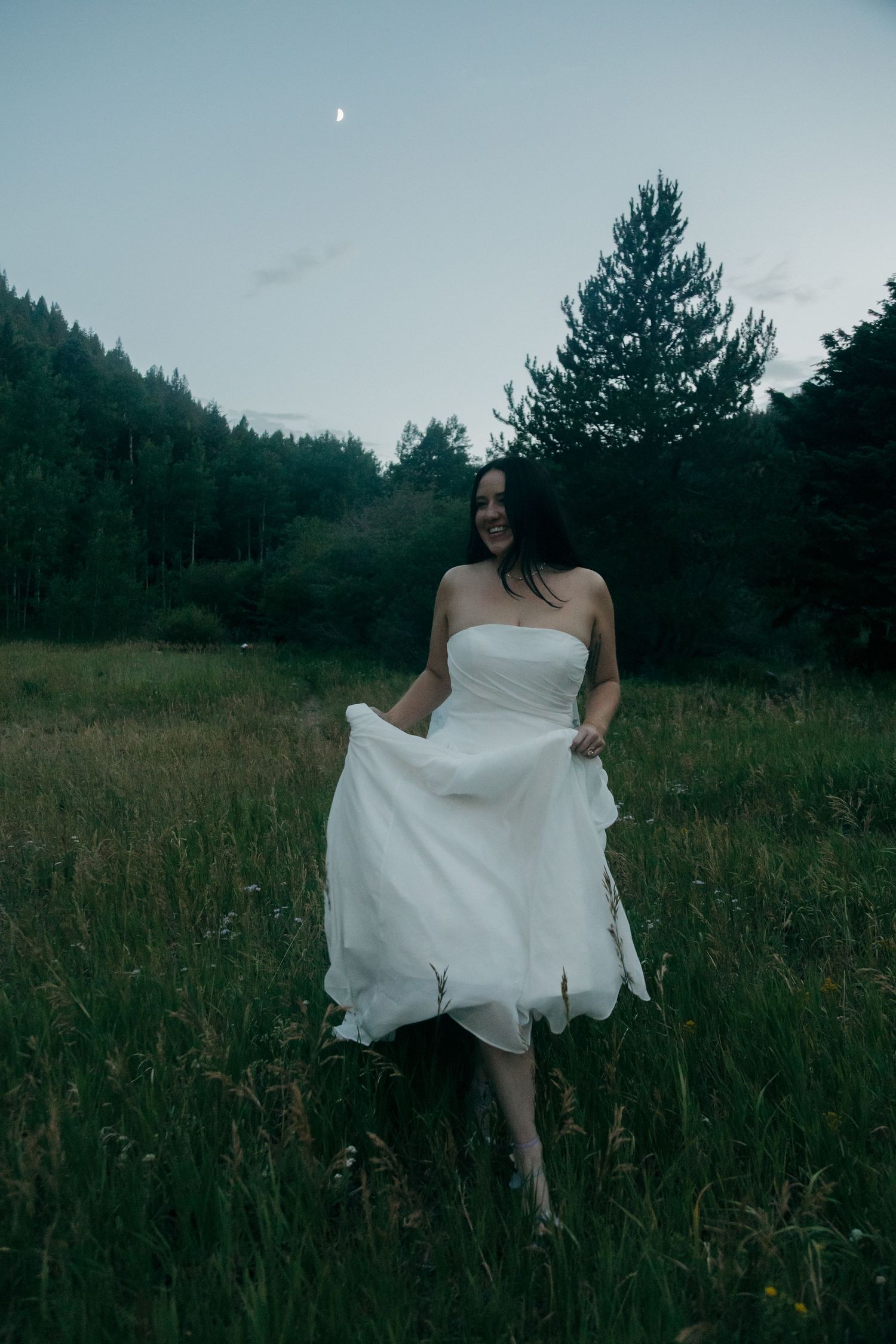 Smiling bride in strapless white gown twirls in green meadow at dusk with crescent moon.