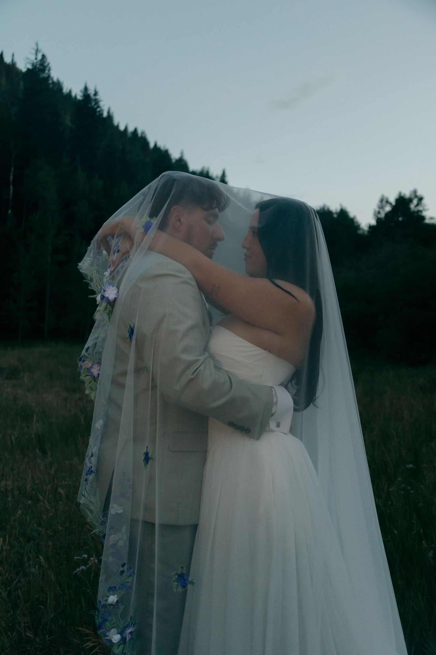 Bride and groom embrace under a floral embroidered veil in a mountain meadow at twilight.
