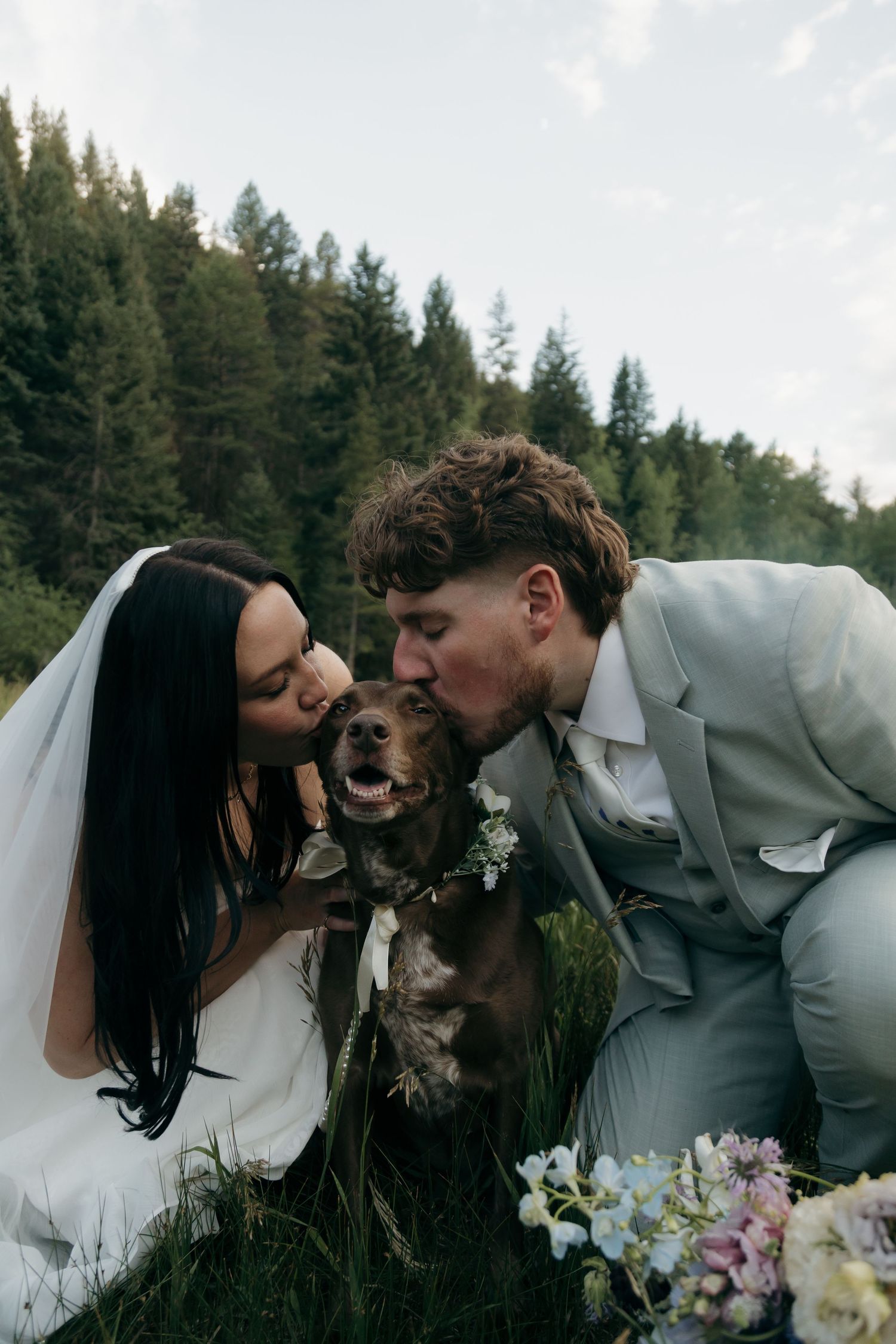 Bride and groom kneel to kiss their brown dog wearing a floral collar at their outdoor mountain wedding.