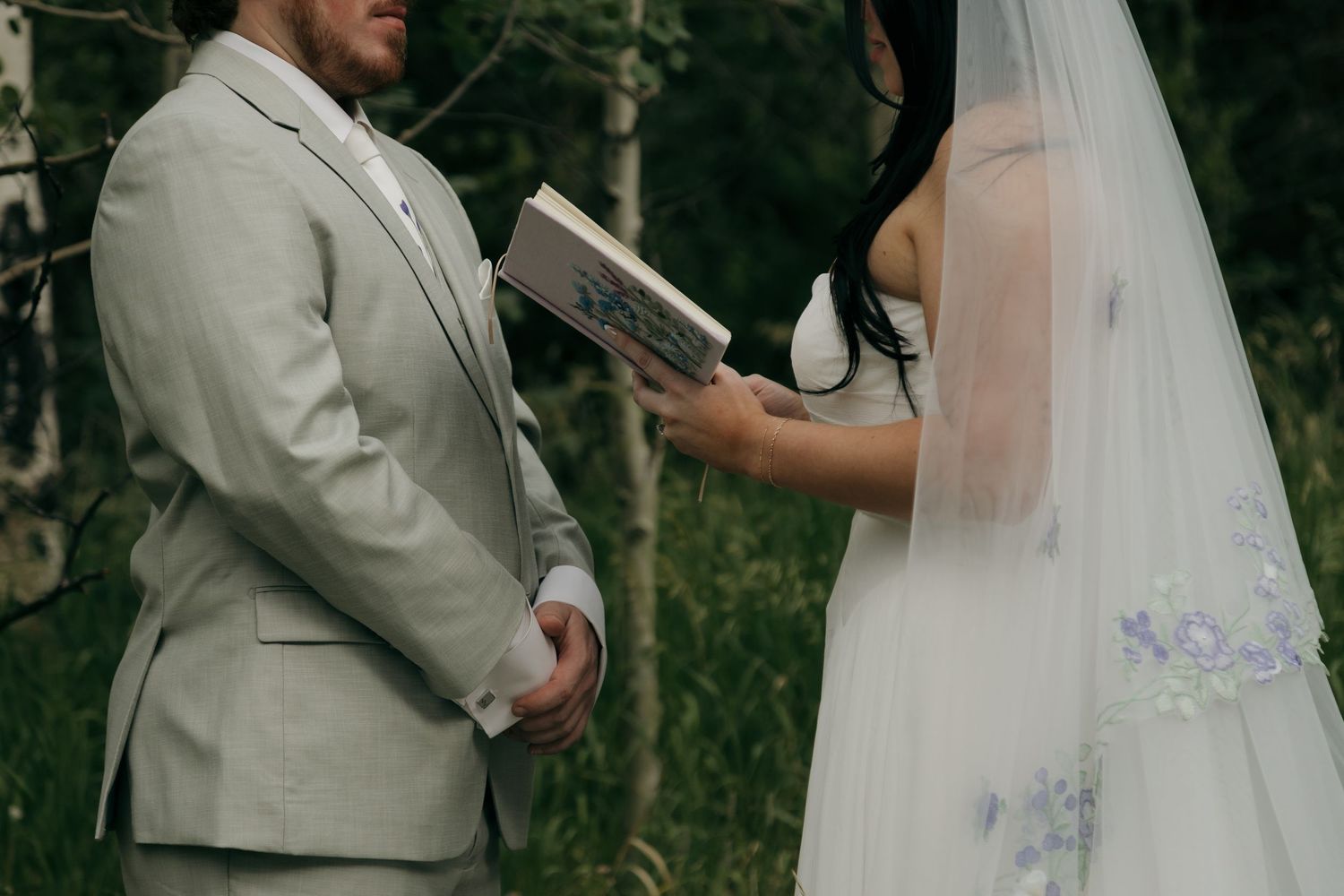 Bride in white floral veil reads vows from a book as groom in light gray suit stands outdoors among trees.