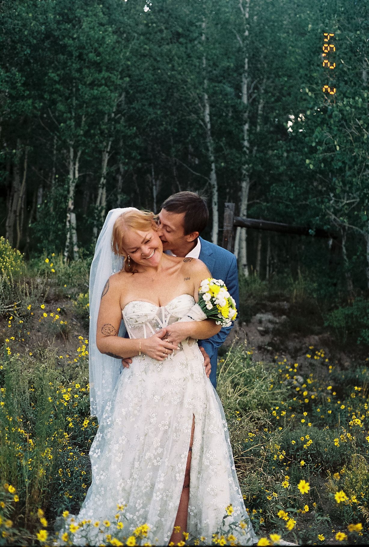 Groom kisses bride on the cheek as she smiles holding a bouquet among wildflowers in an aspen forest.