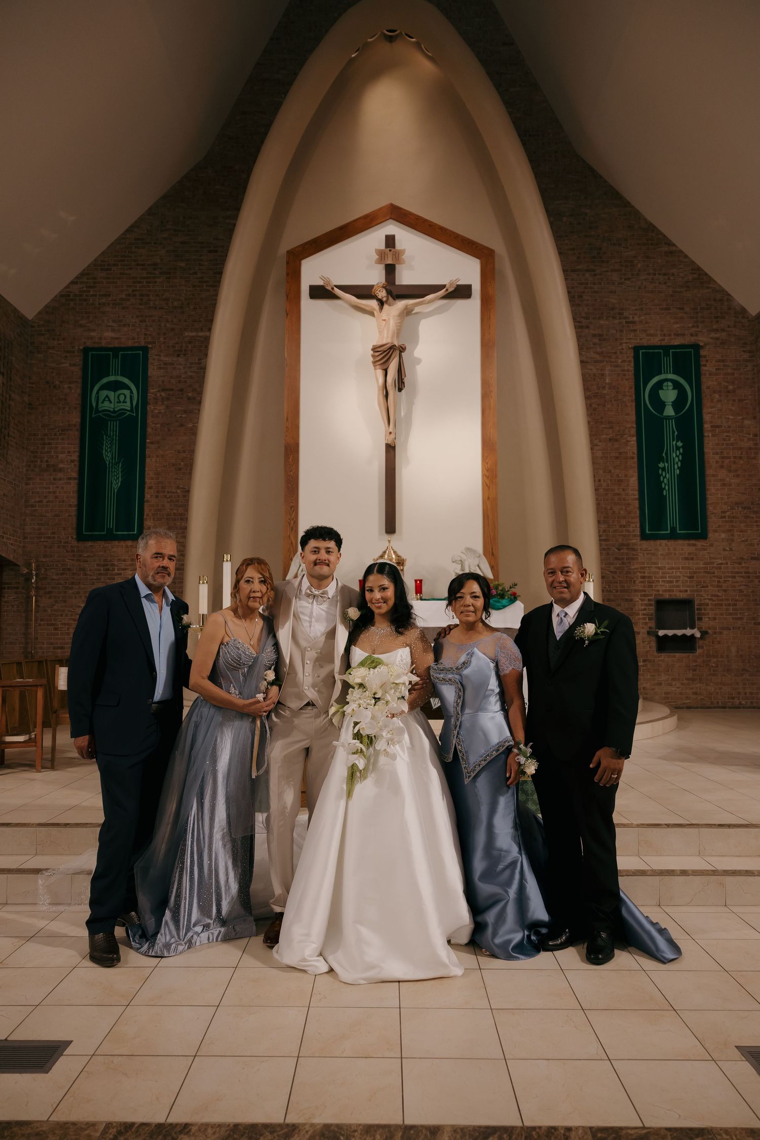 Wedding party poses before altar with crucifix in Catholic church, bride in white gown with groom in tan suit flanked by family.