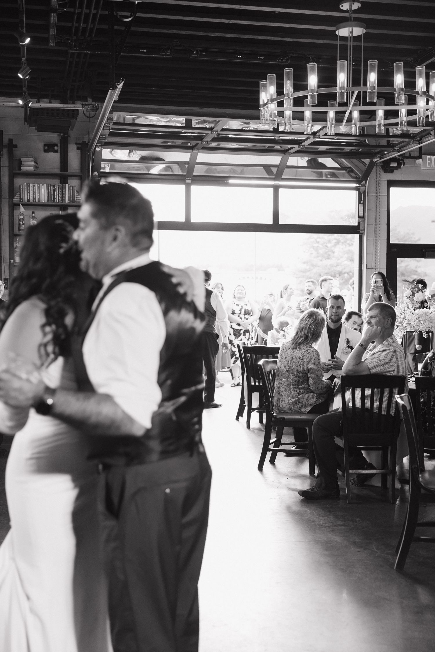 Black and white photo of couple sharing first dance while guests watch at reception.