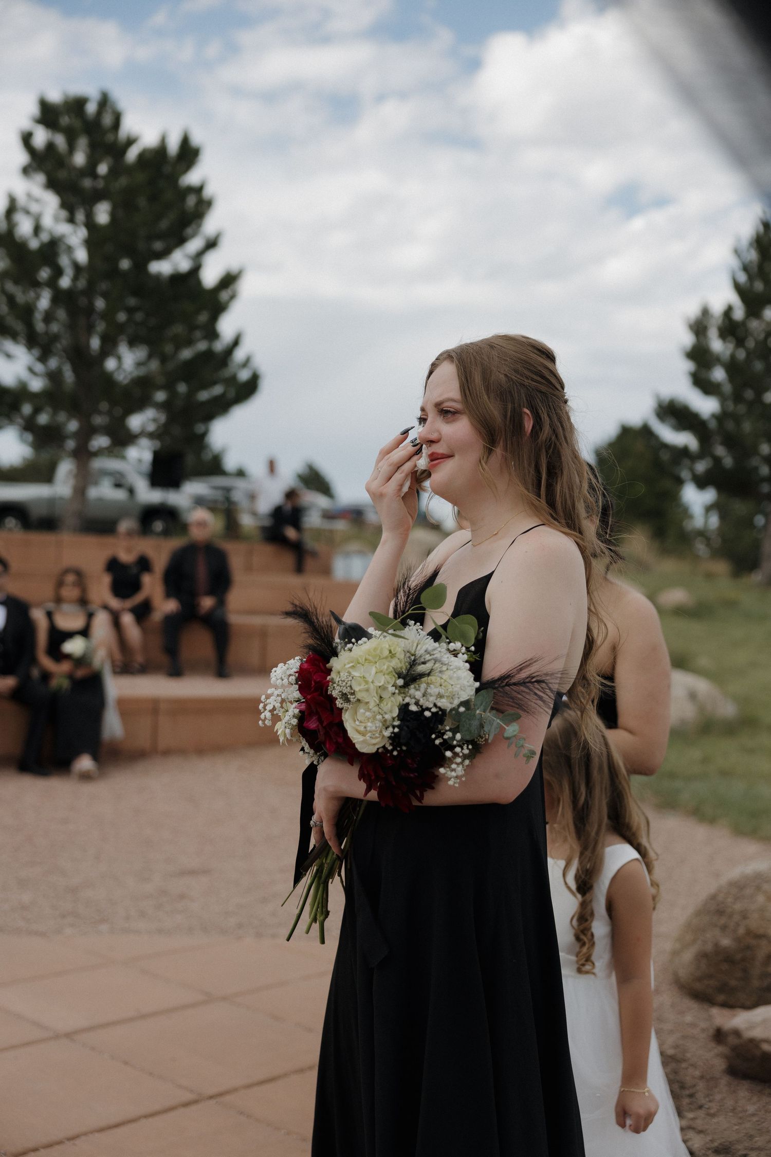 Emotional bridesmaid in black dress wipes tears while holding bouquet during outdoor wedding ceremony.