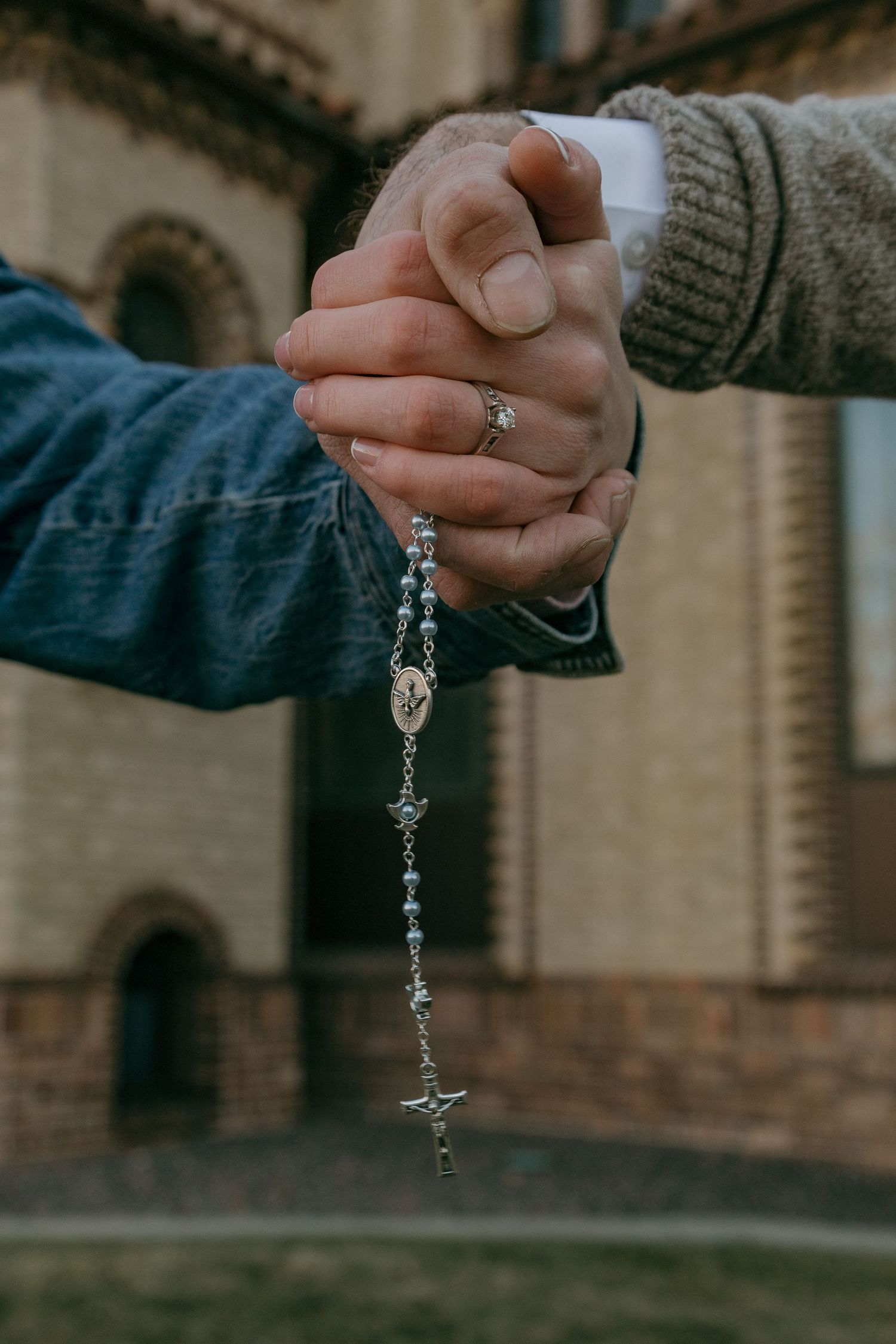 A close-up series of hands holding a delicate silver rosary against a blurred architectural background.