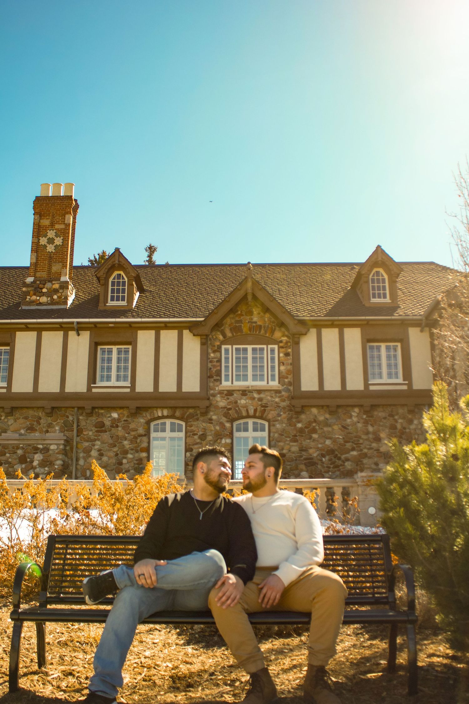 A loving couple sits together on a park bench outside a stone mansion with Tudor architectural details.