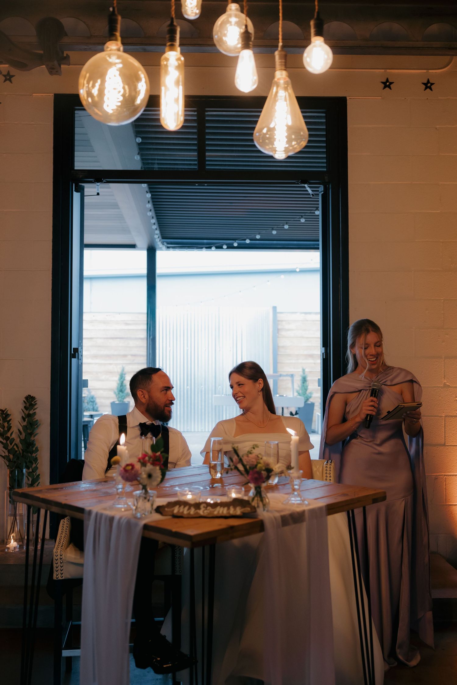 Bride and groom seated at sweetheart table laughing as bridesmaid gives speech at wedding reception.