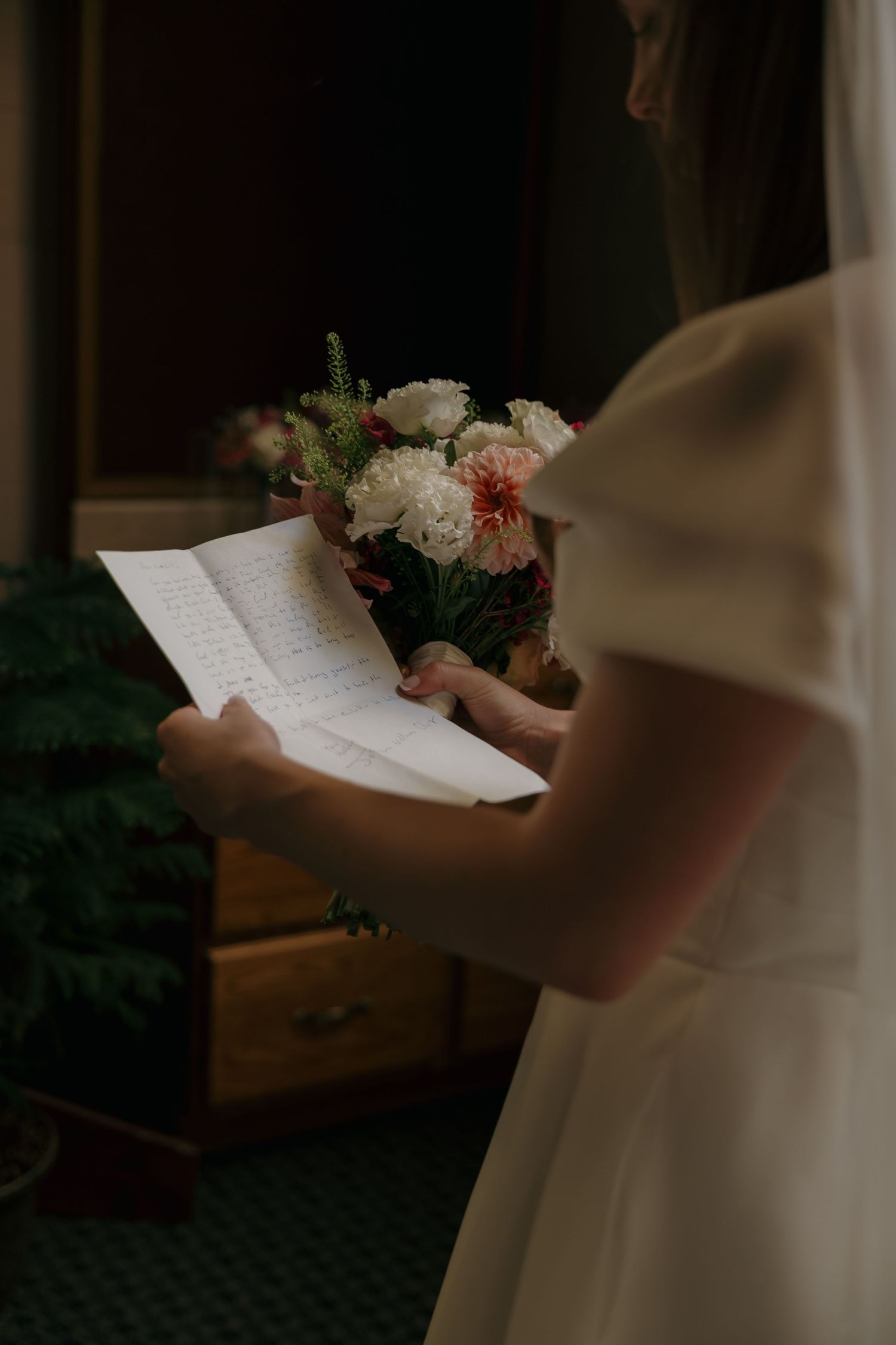Bride in white dress reading handwritten vows while holding a bouquet of white and pink flowers before her wedding ceremony.