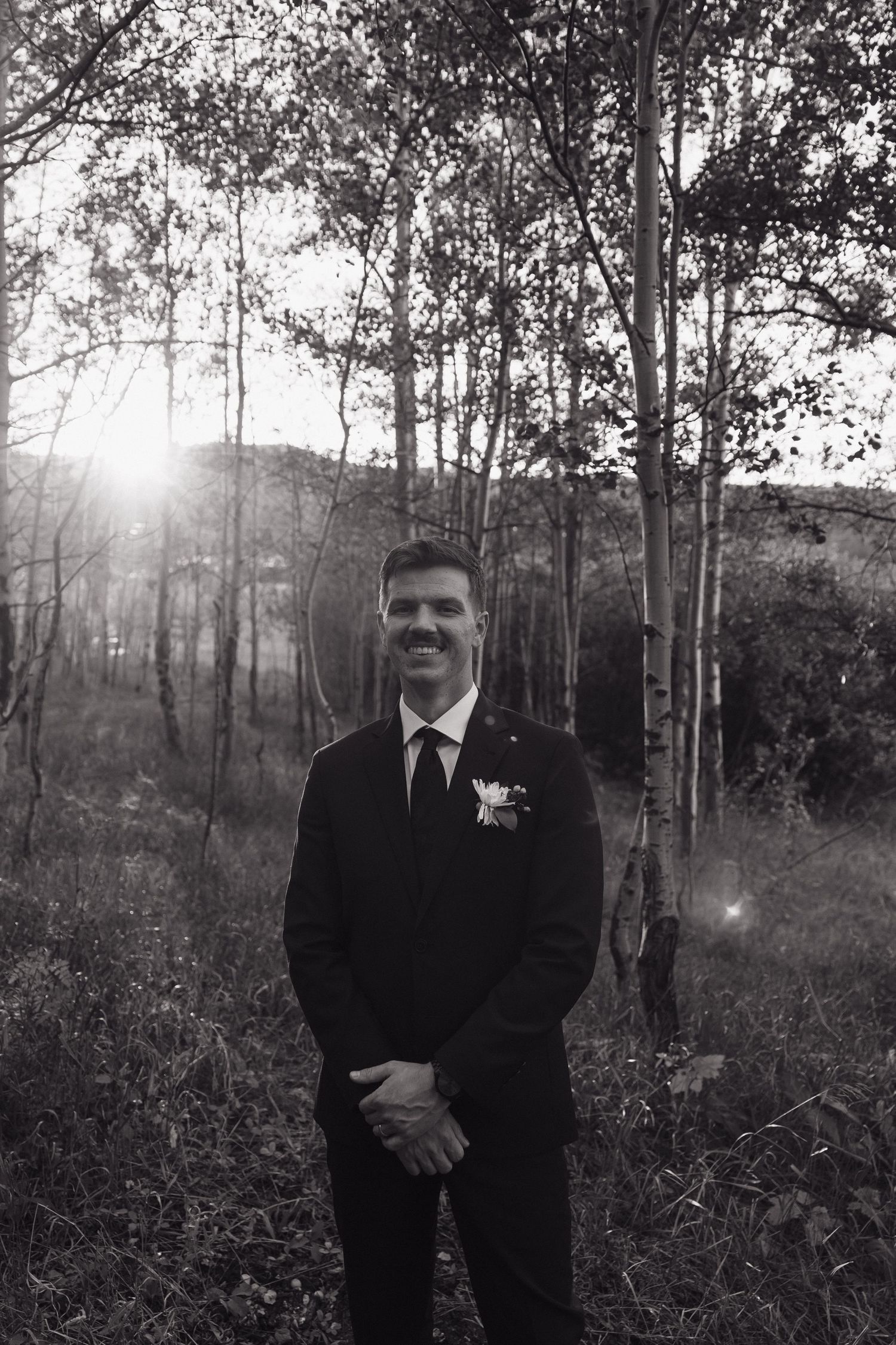 Black and white portrait of smiling groom in dark suit with boutonniere standing among aspen trees.