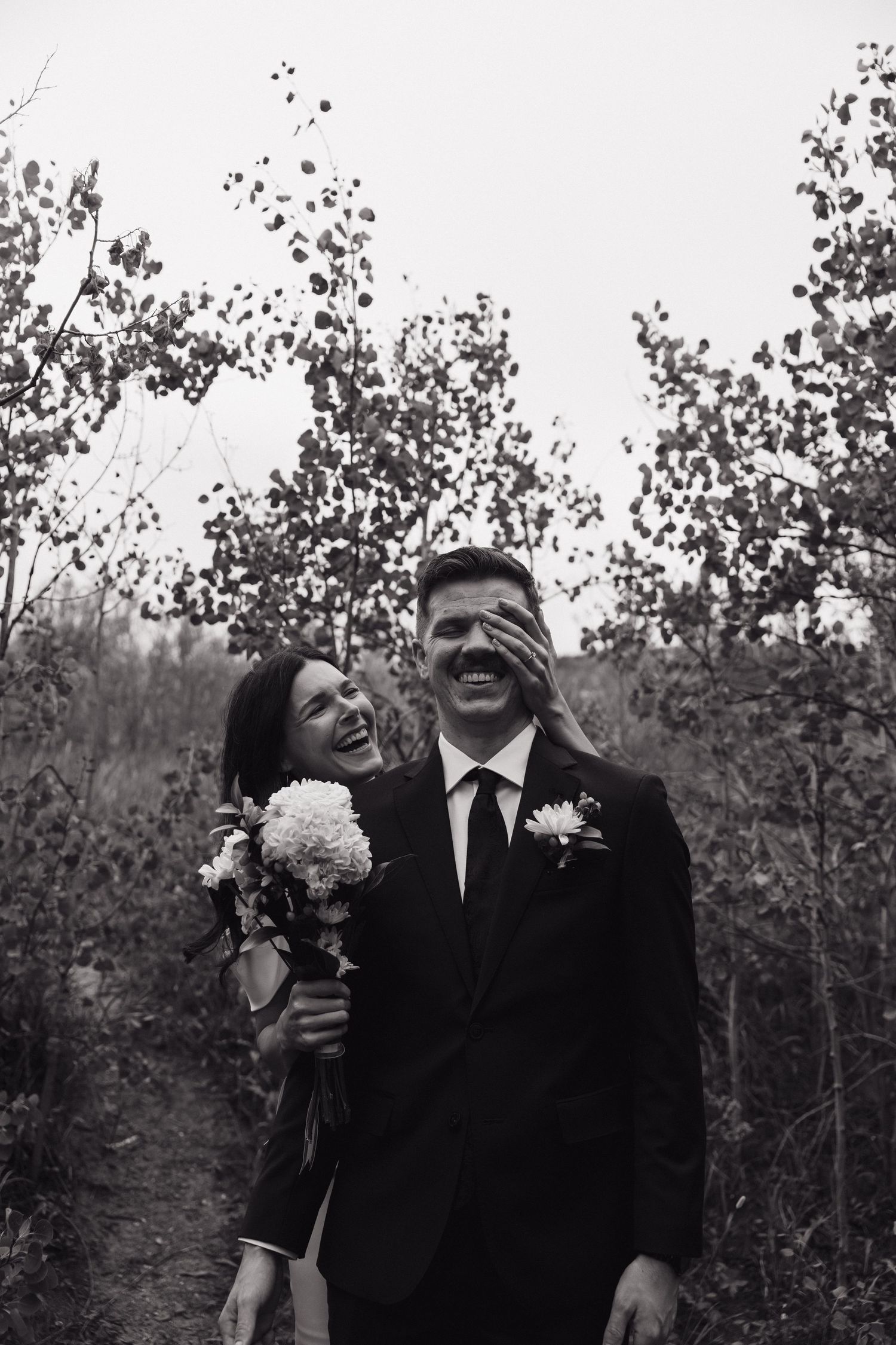 Black and white photo of laughing bride covering grooms eyes in autumn forest.