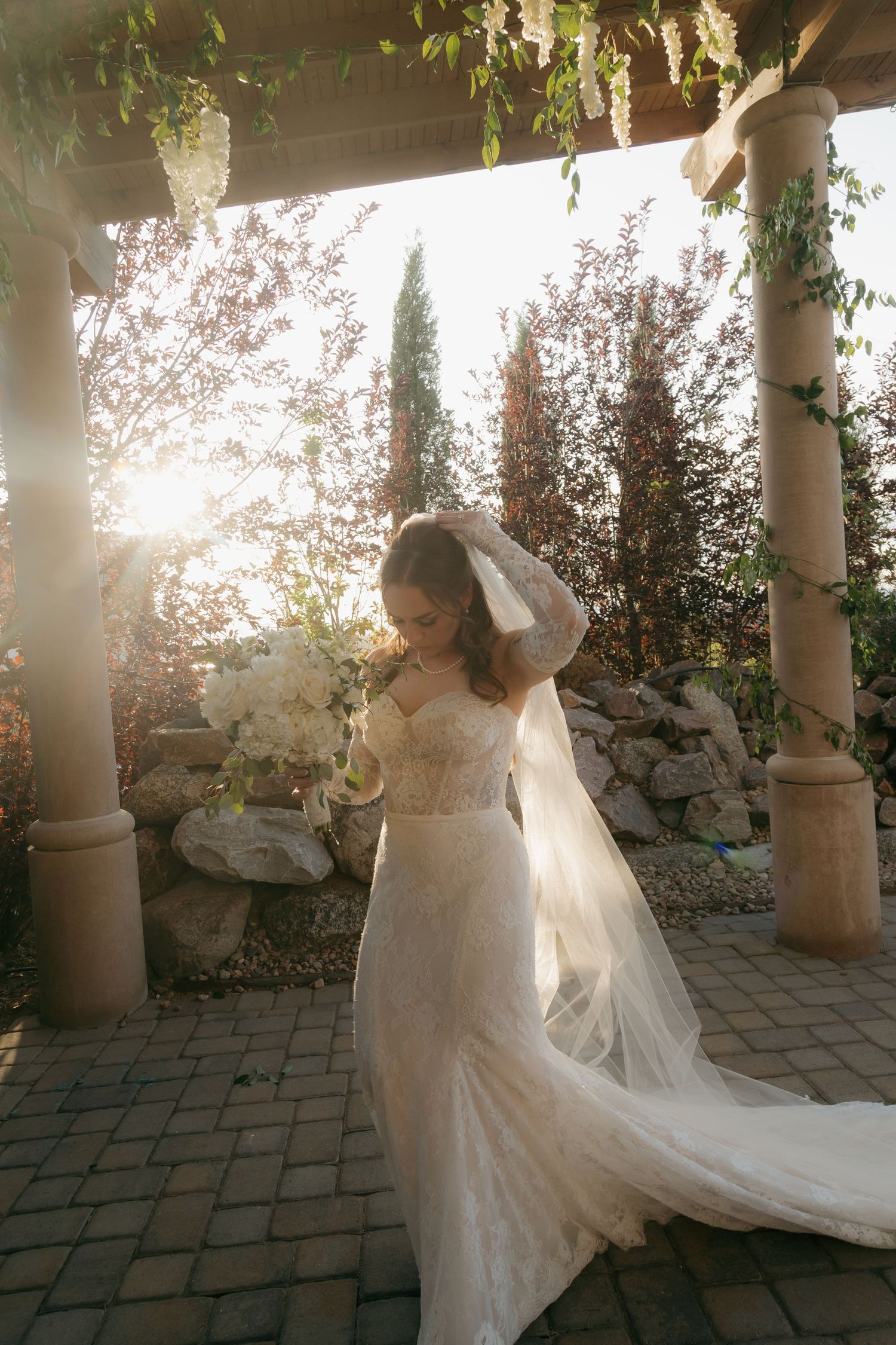 Bride in lace wedding gown and veil posing under pergola with sunlight streaming through garden.