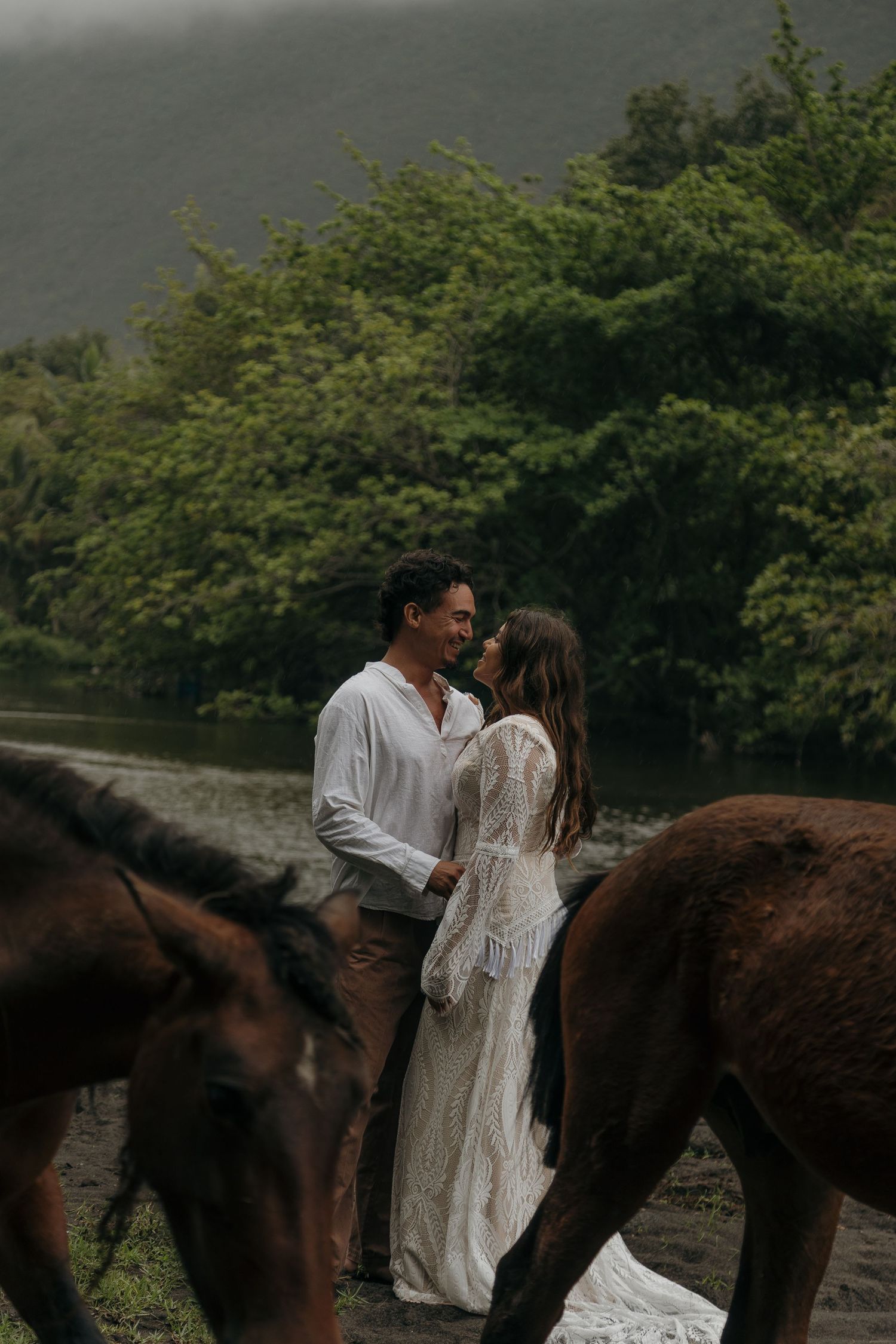 A couple in white outfits stands together with horses by a river surrounded by lush greenery.