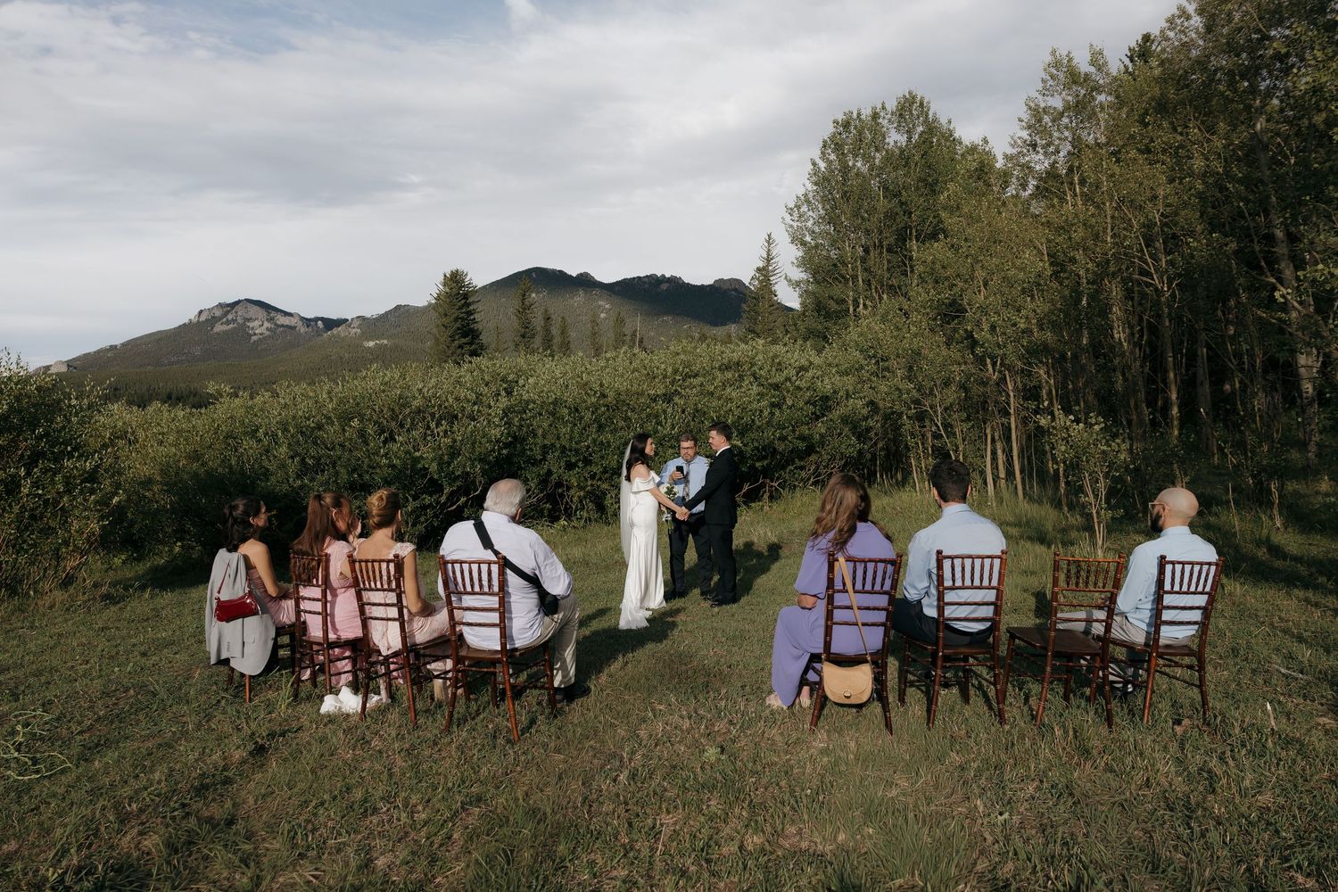 Wide shot of small outdoor wedding ceremony with guests seated in wooden chairs amid mountains.