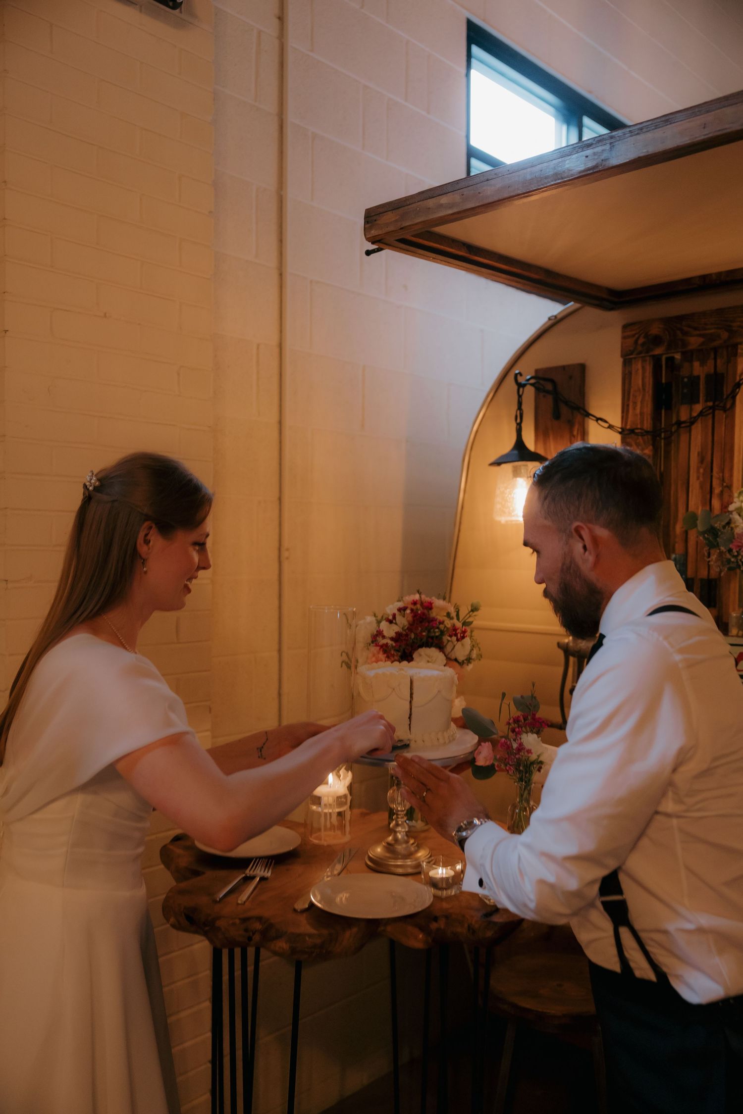 Bride and groom cutting wedding cake together at rustic wooden table with candles in warmly lit venue.