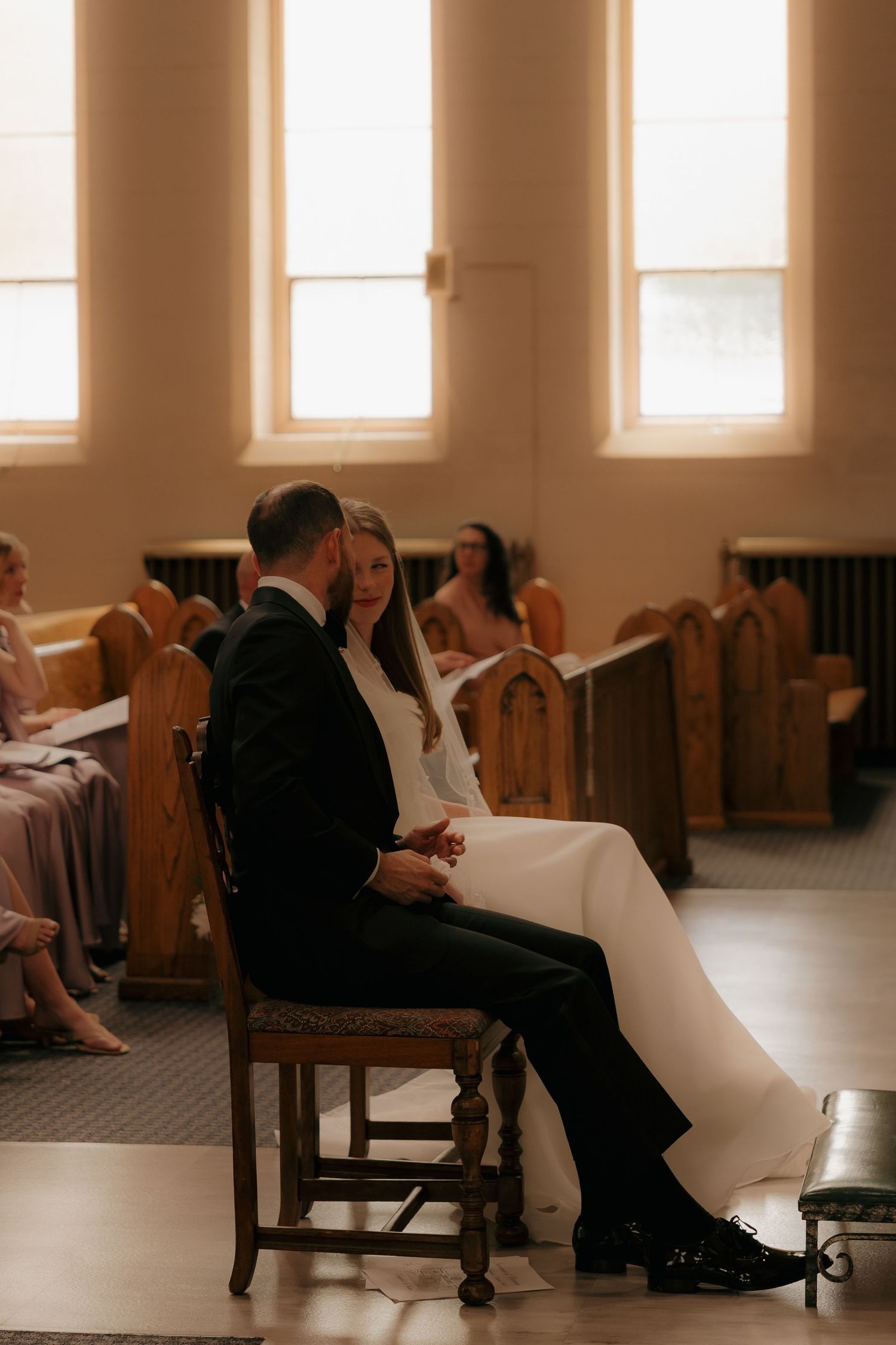 Bride and groom sit together holding hands during wedding ceremony in a sunlit church.