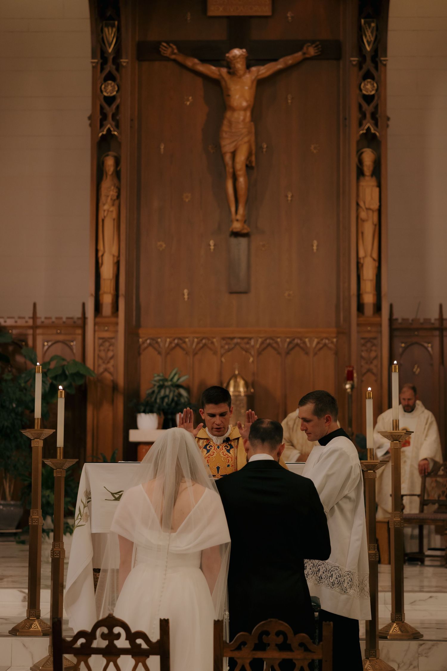 Bride and groom kneel before priest at ornate wooden altar with crucifix during Catholic wedding mass.