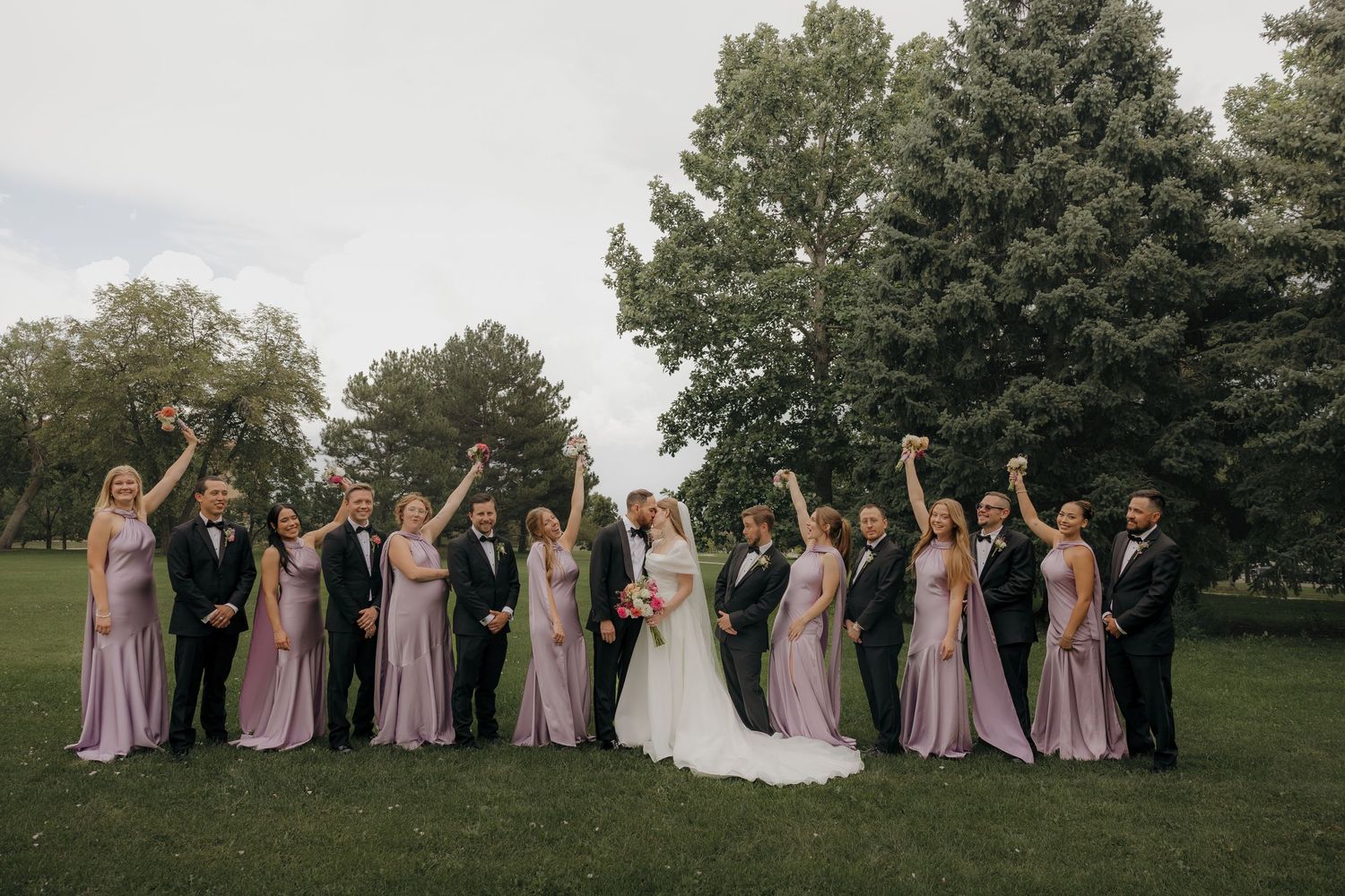 Wedding party in lavender dresses and black suits celebrating outdoors with arms raised.