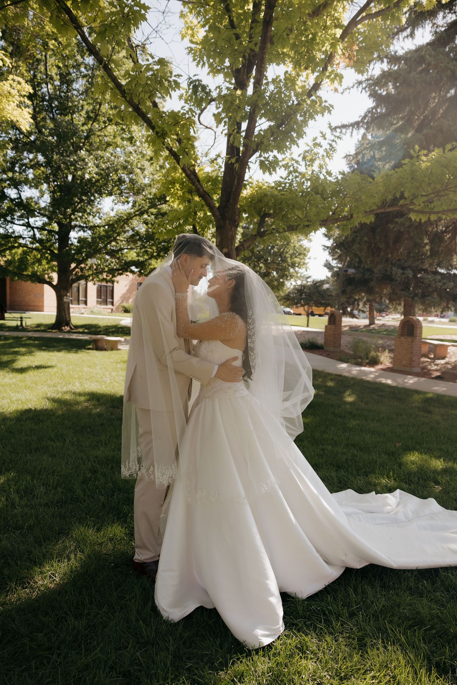 Bride and groom sharing romantic kiss under veil in sunlit garden after wedding ceremony.