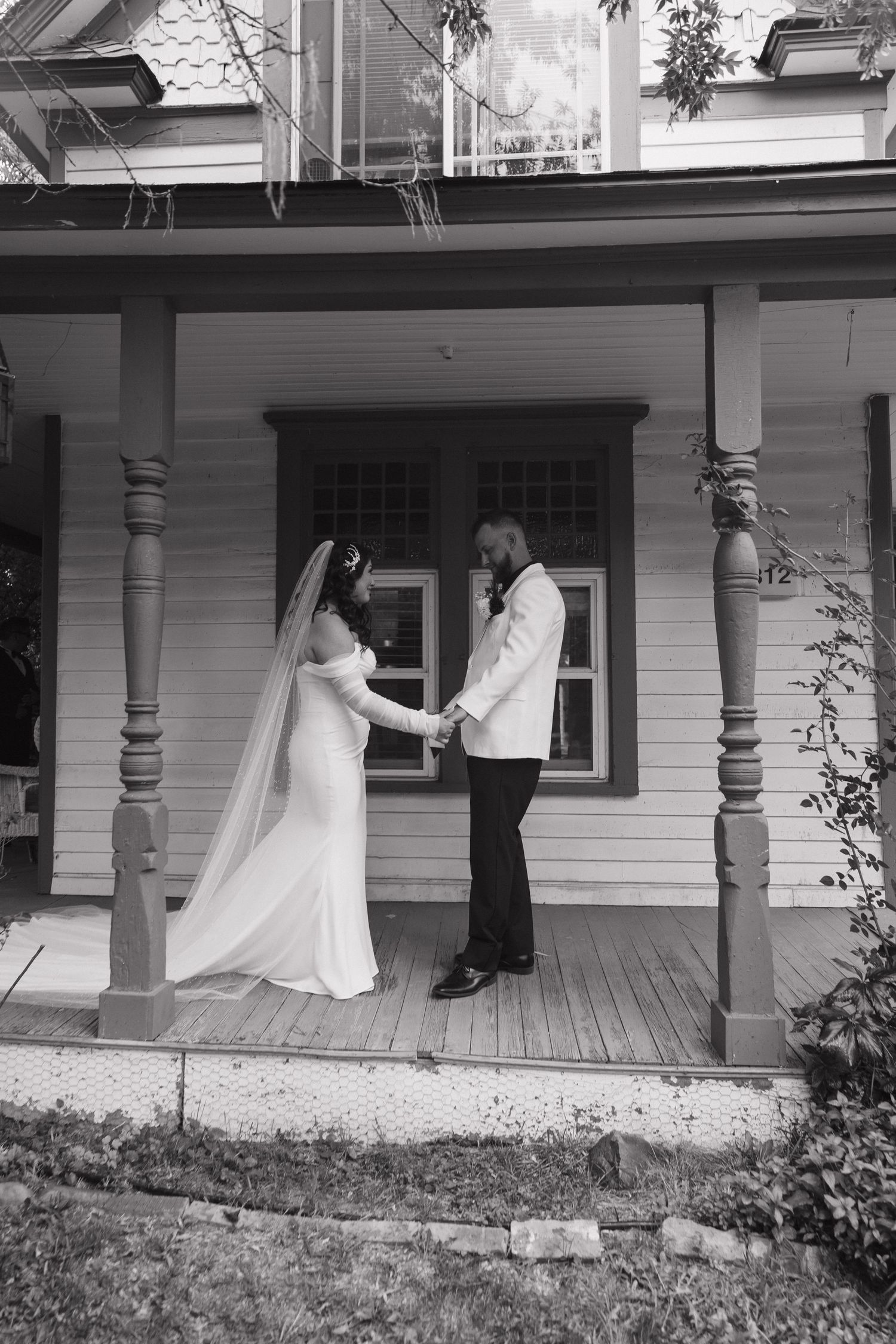 Black and white photo of bride and groom holding hands on a vintage home porch on their wedding day.