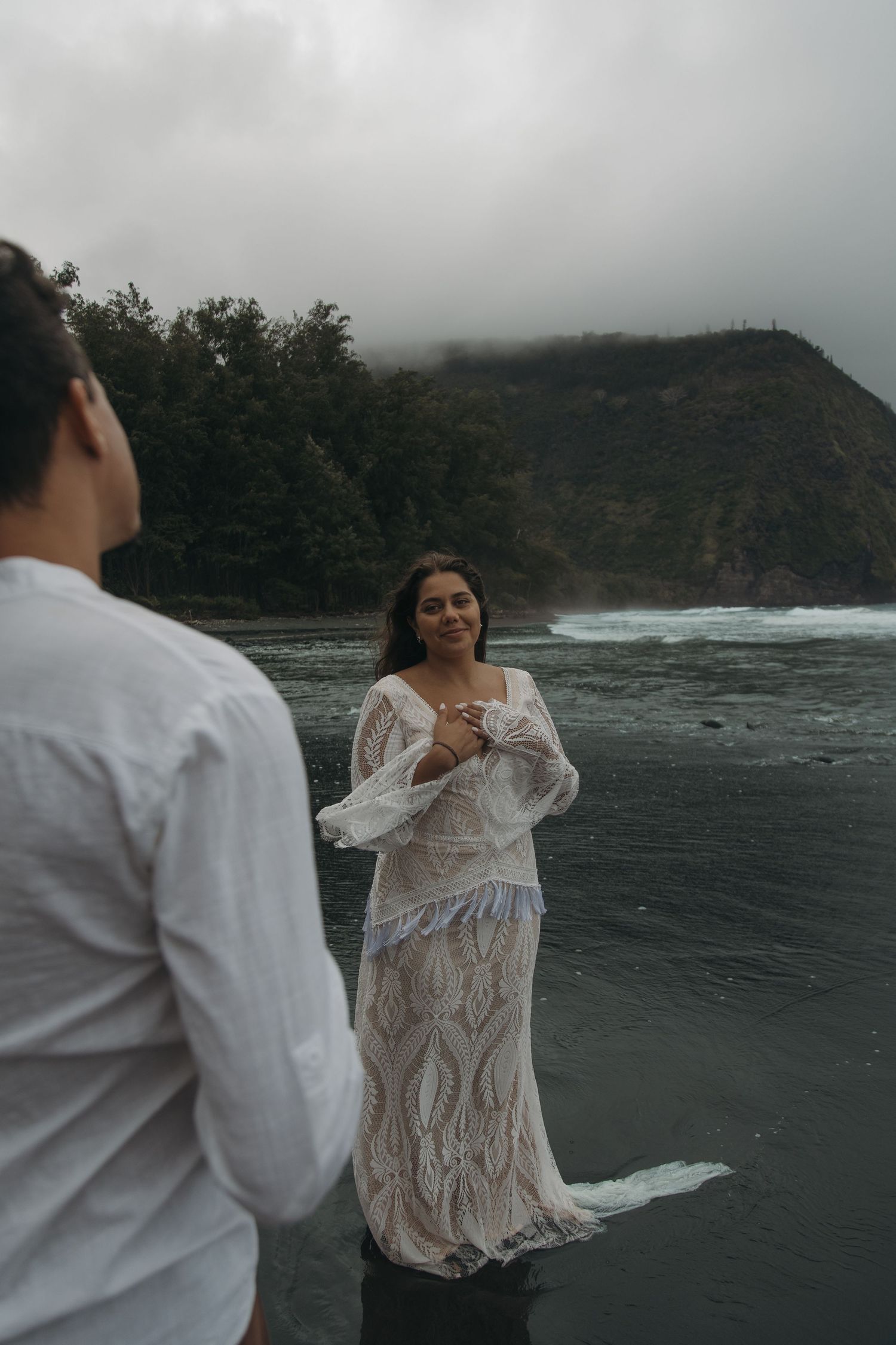 Bride in bohemian lace gown smiles at groom on misty black sand beach with mountain backdrop.