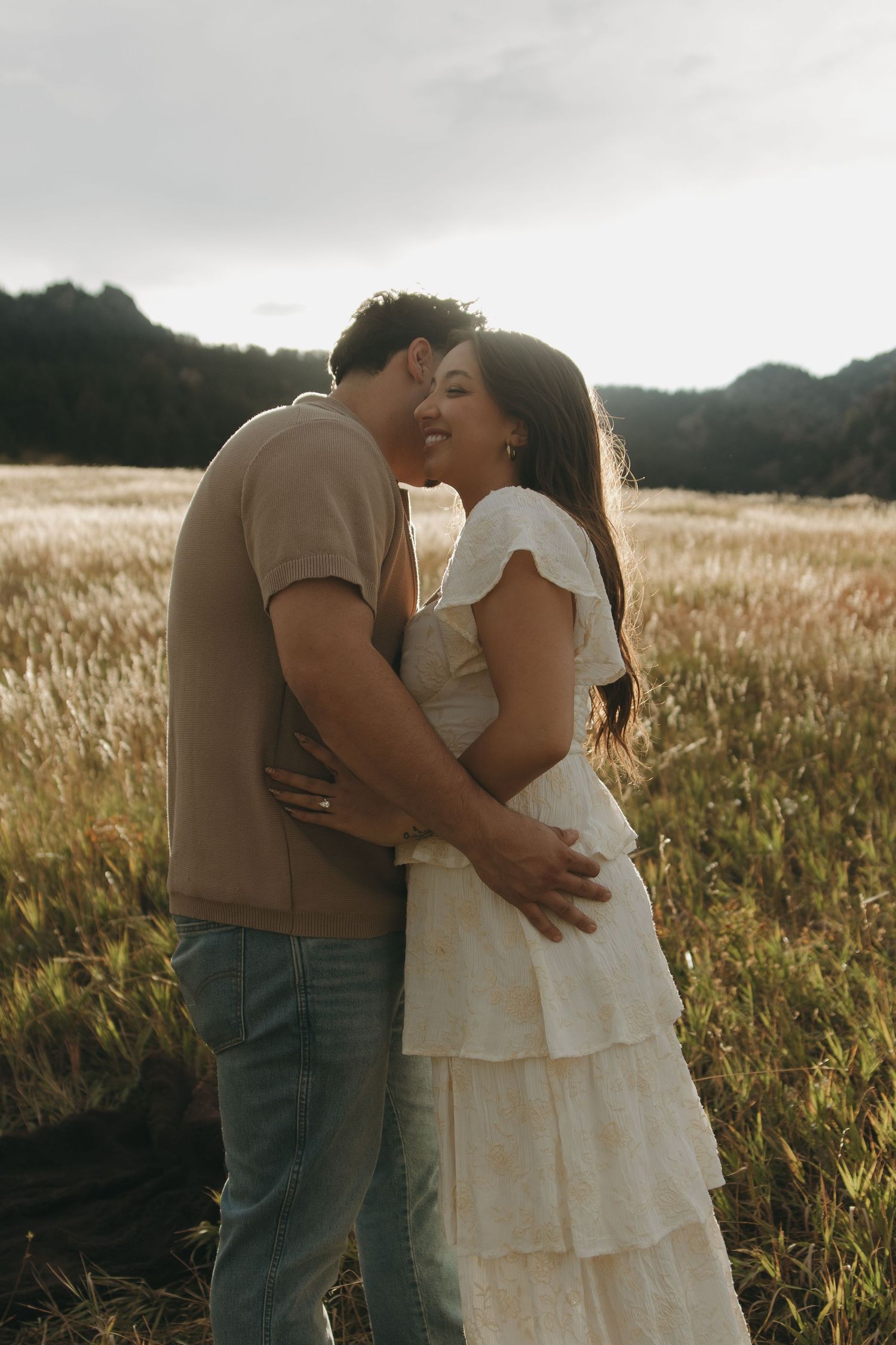 Man kissing smiling woman in white ruffled dress in a golden field at sunset.