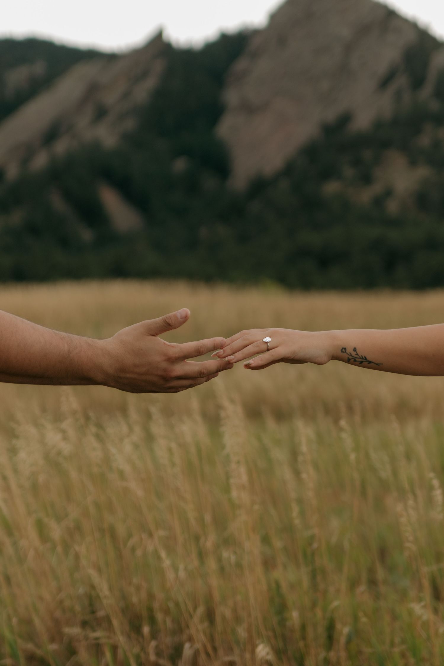 Close-up of couple reaching hands toward each other showing engagement ring with Flatirons mountains behind.