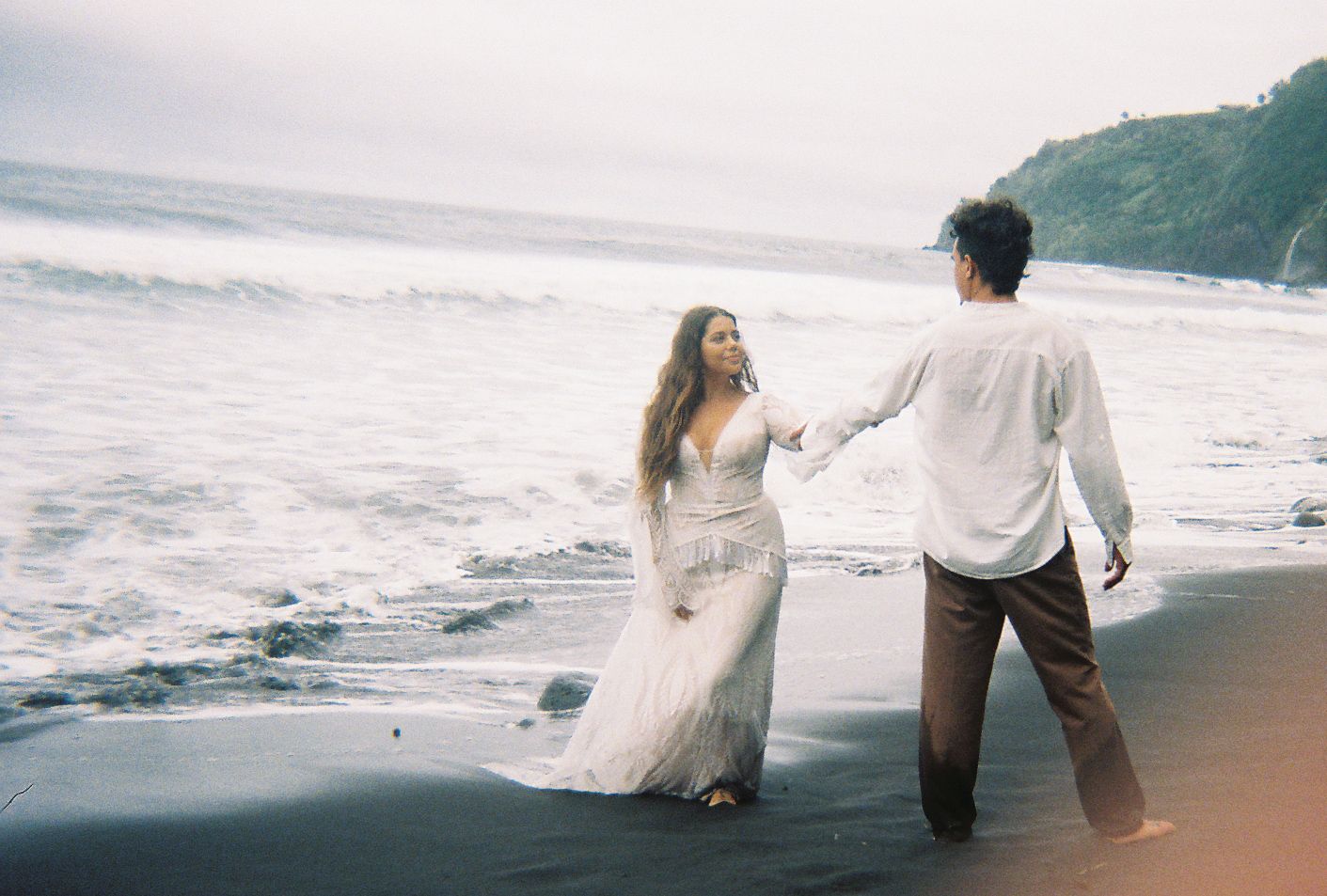 Bride and groom holding hands on black sand beach with ocean waves in Hawaii.