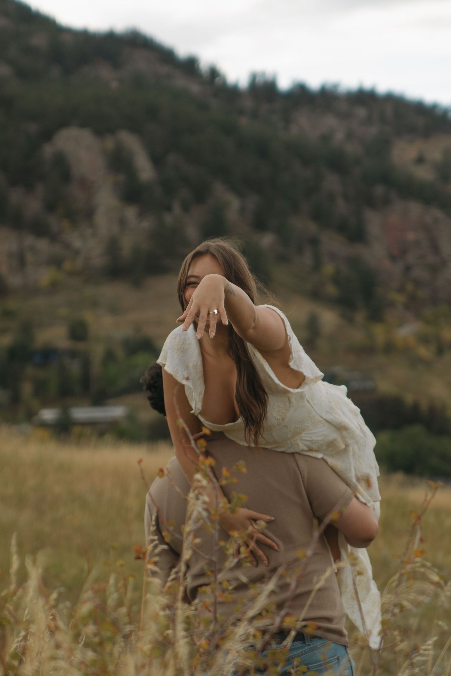 Man lifts woman in white dress in golden meadow with mountain forest backdrop.