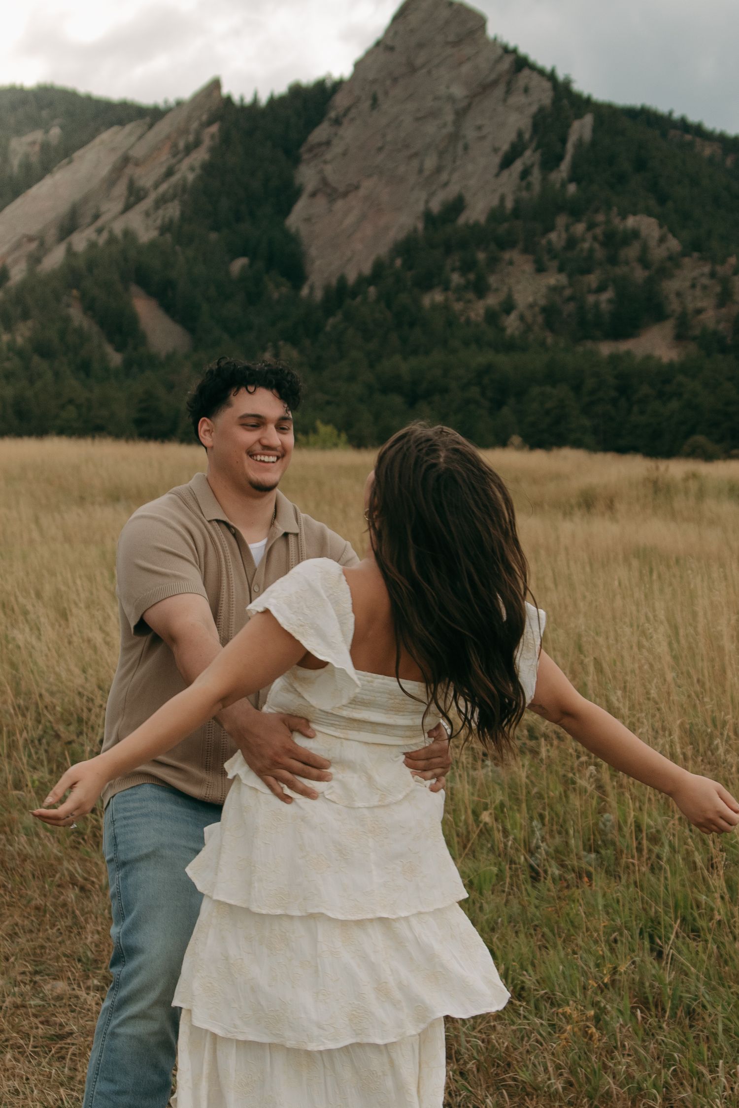 Man smiling while dancing with woman in white tiered dress in a mountain meadow.