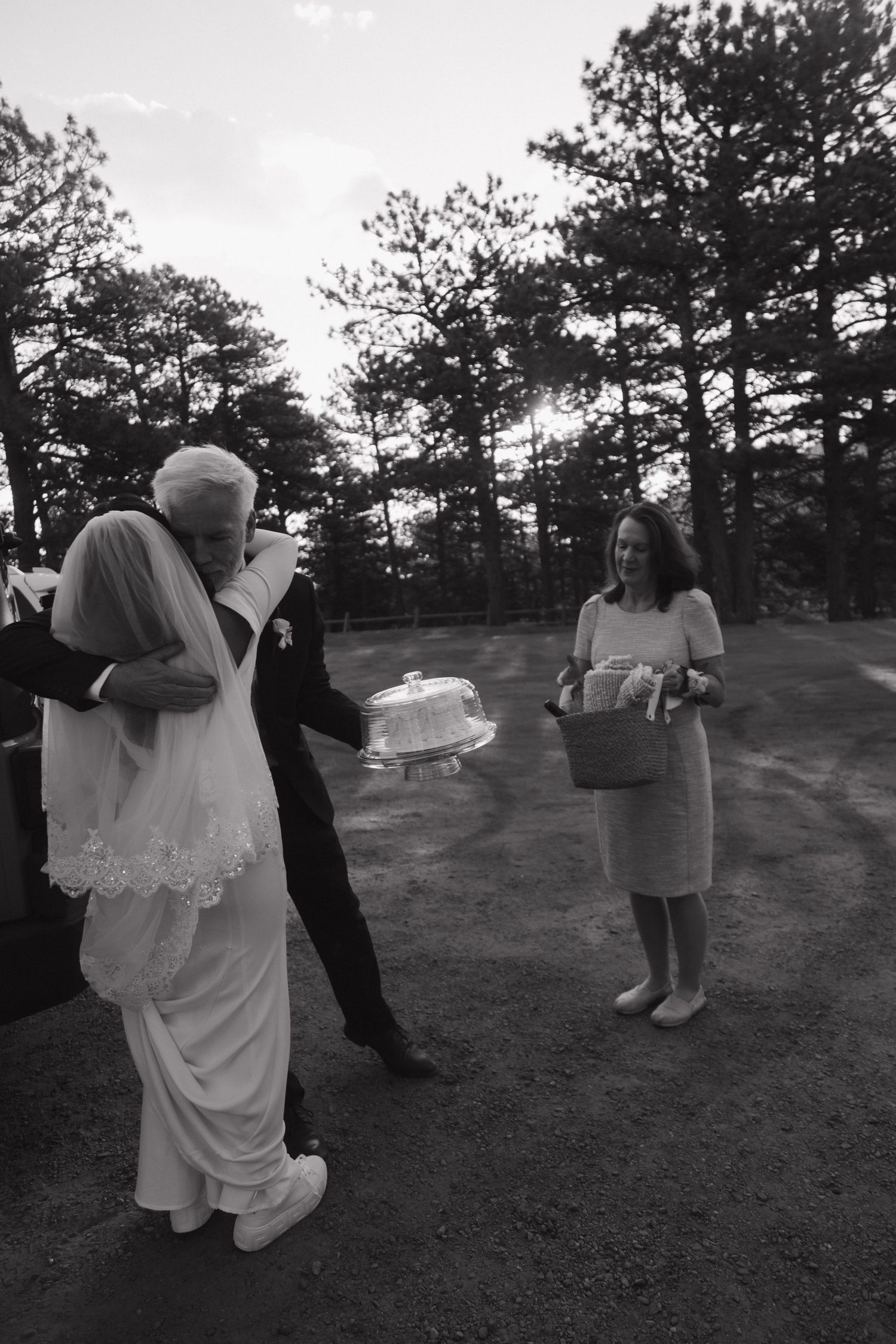 Black and white photo of bride and groom receiving wedding cake outdoors among pine trees.